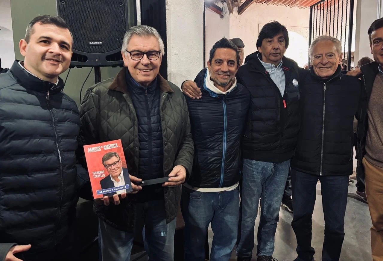 Grupo de seis hombres en una reunión, uno sostiene un libro titulado 'Amigos de América' con una foto de un hombre en la portada