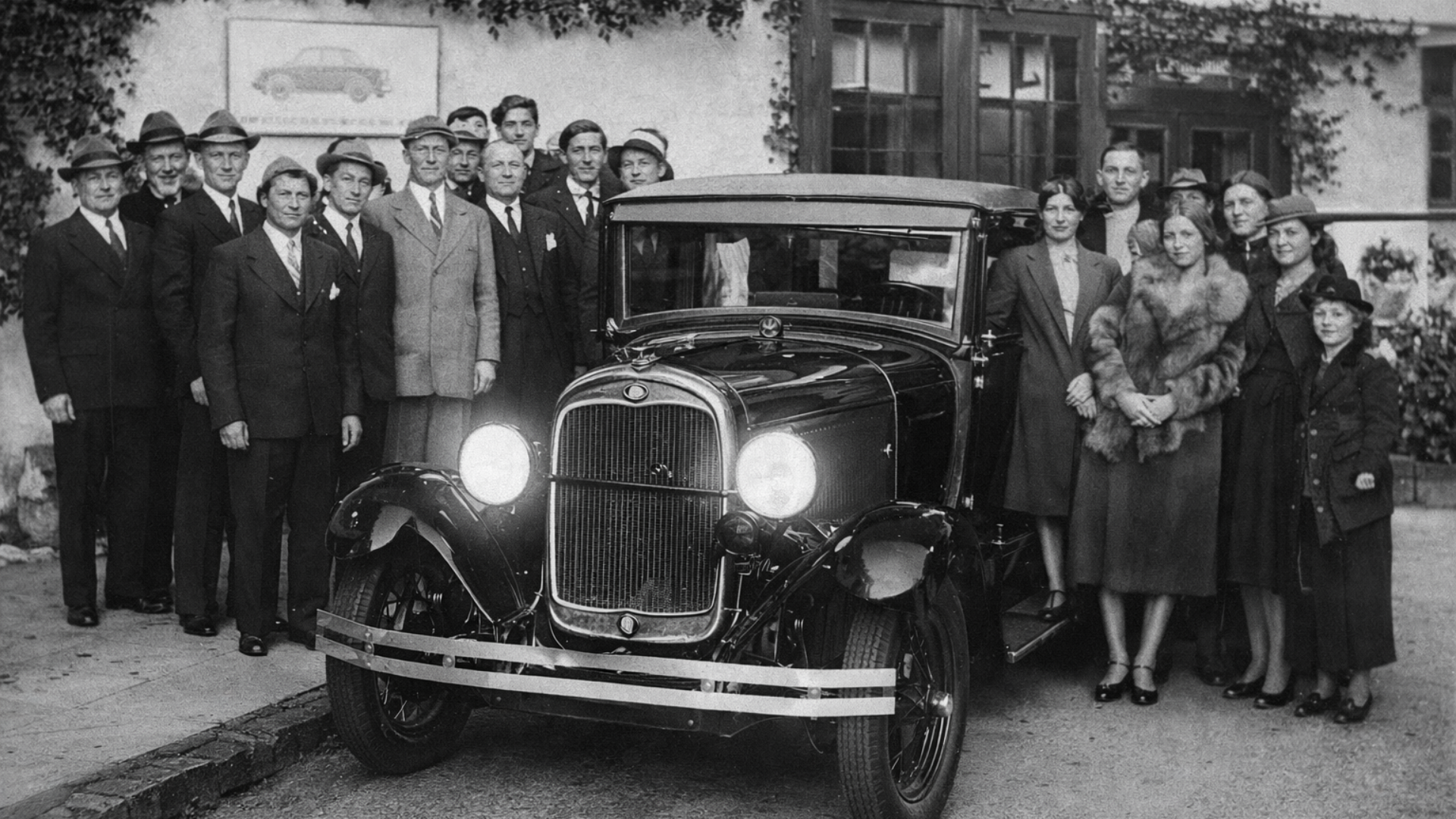 Grupo de personas posando con un automóvil clásico frente a una casa antigua rodeada de plantas.