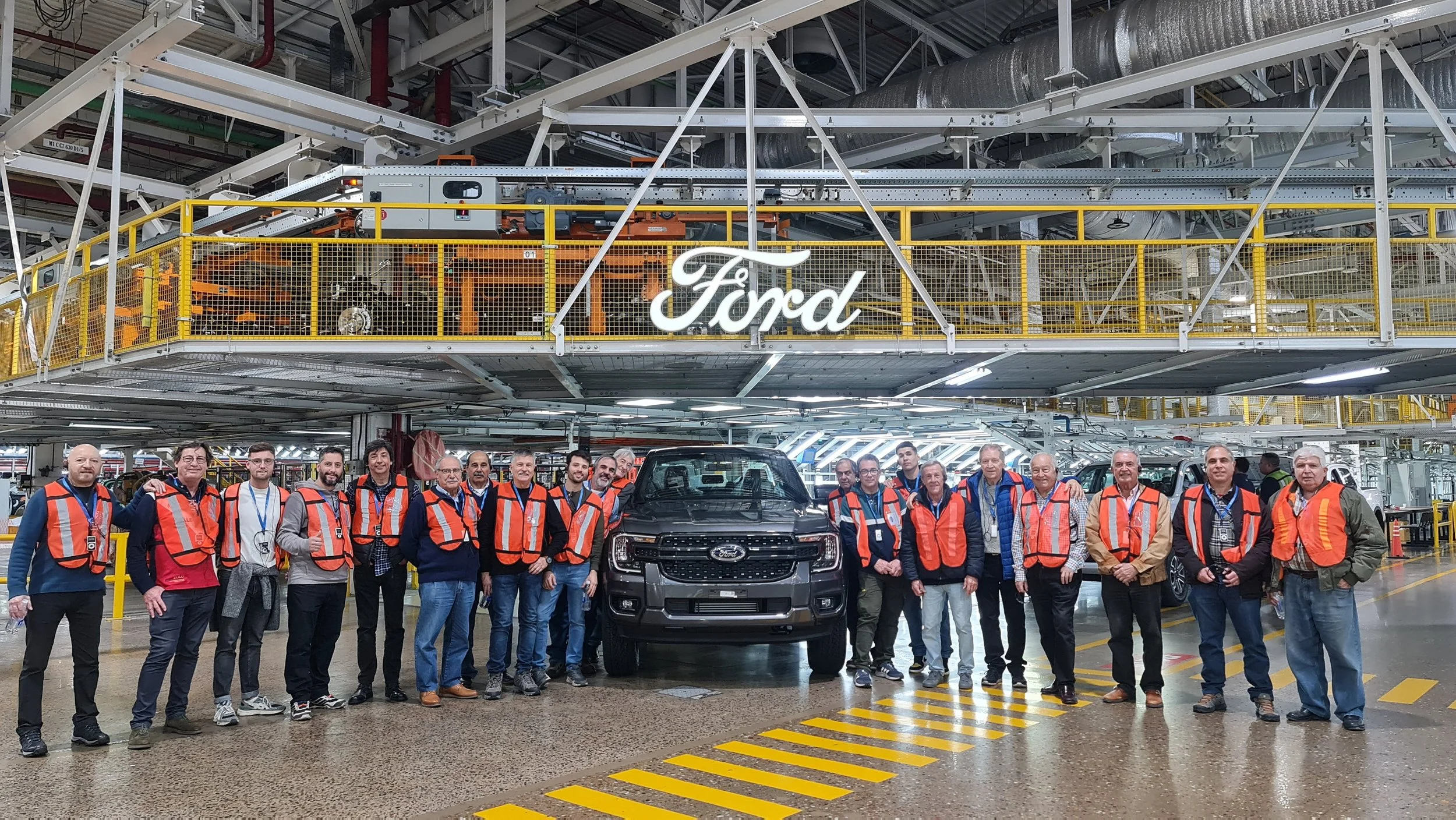 Grupo de personas en una fábrica de vehículos Ford, algunas con chalecos de seguridad naranjas, rodeando una camioneta Ford en el interior de la planta de ensamblaje.
