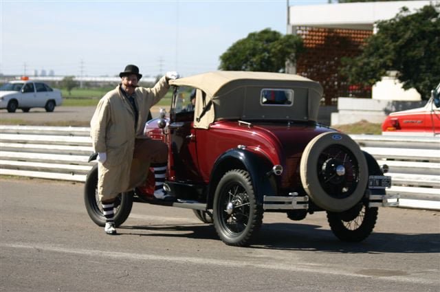 Hombre vestido con ropa antigua junto a un auto clásico rojo con capota beige y neumático de repuesto en la parte trasera, en una vía de carreras.