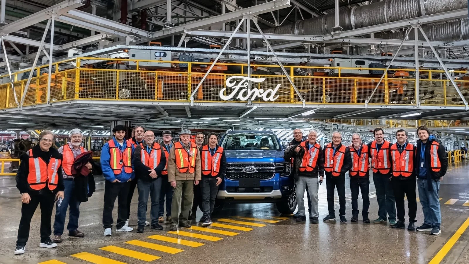 Grupo de personas con chalecos de seguridad posando frente a una camioneta en una planta de fabricación de Ford.