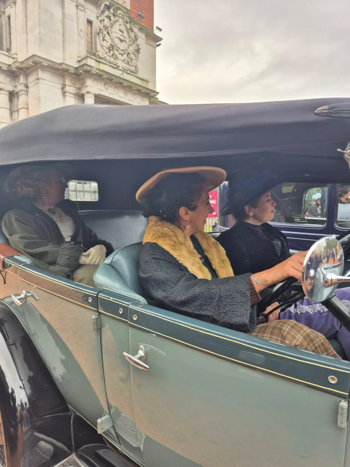 Tres mujeres vestidas con ropa vintage y sombreros, viajando en un coche antiguo, con un edificio histórico al fondo.