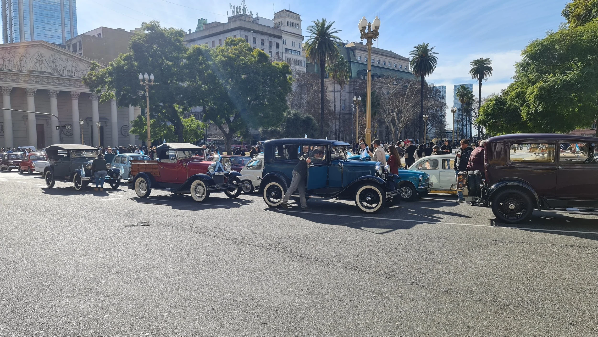 Auto antiguos en una calle concurrida con gente y árboles, en un entorno urbano con edificios altos y palmeras.