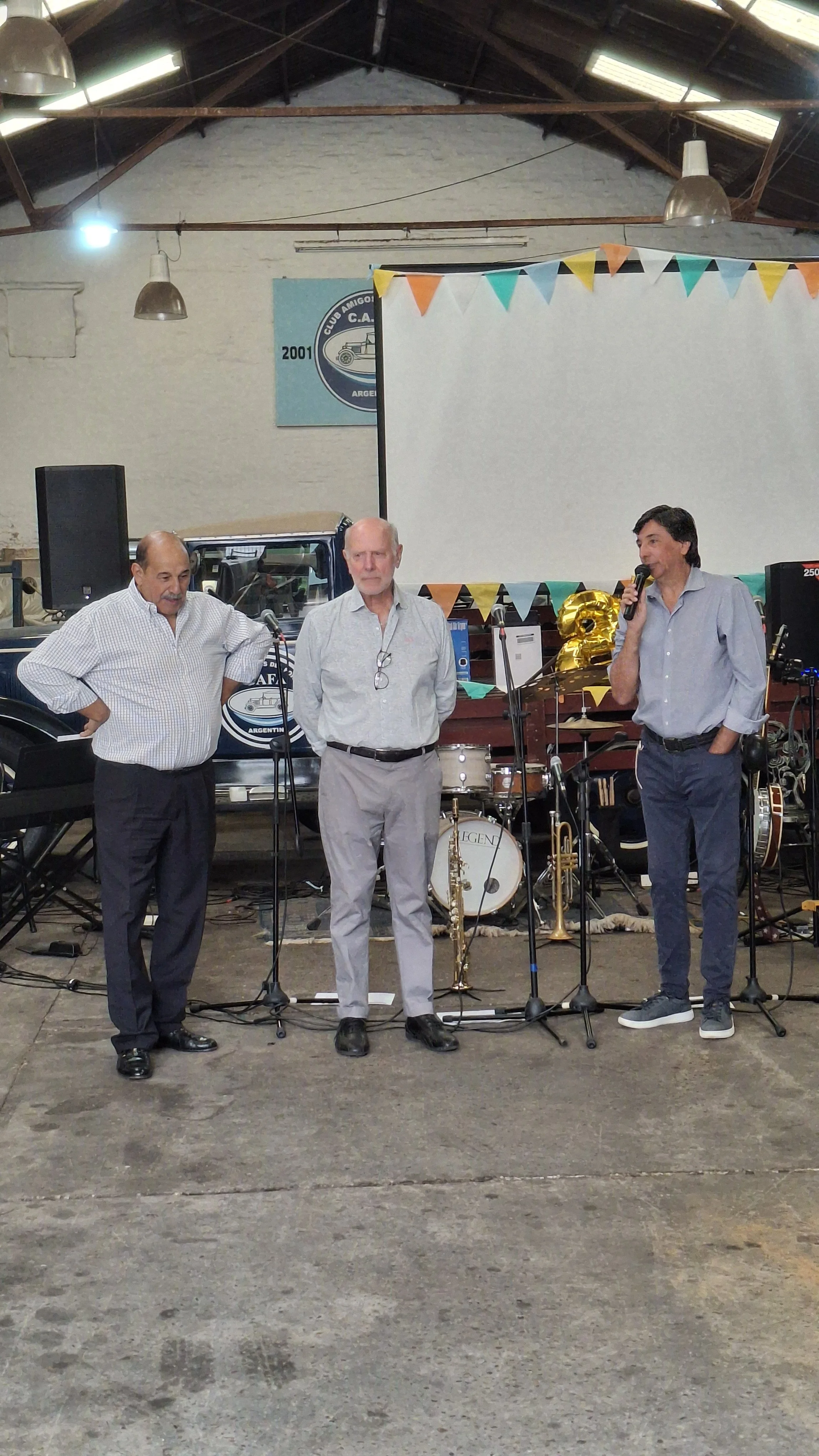 Tres hombres en un escenario con instrumentos musicales y decoraciones de colores, una pantalla blanca y un cartel con el logo de un club de autos antiguo en Argentina.