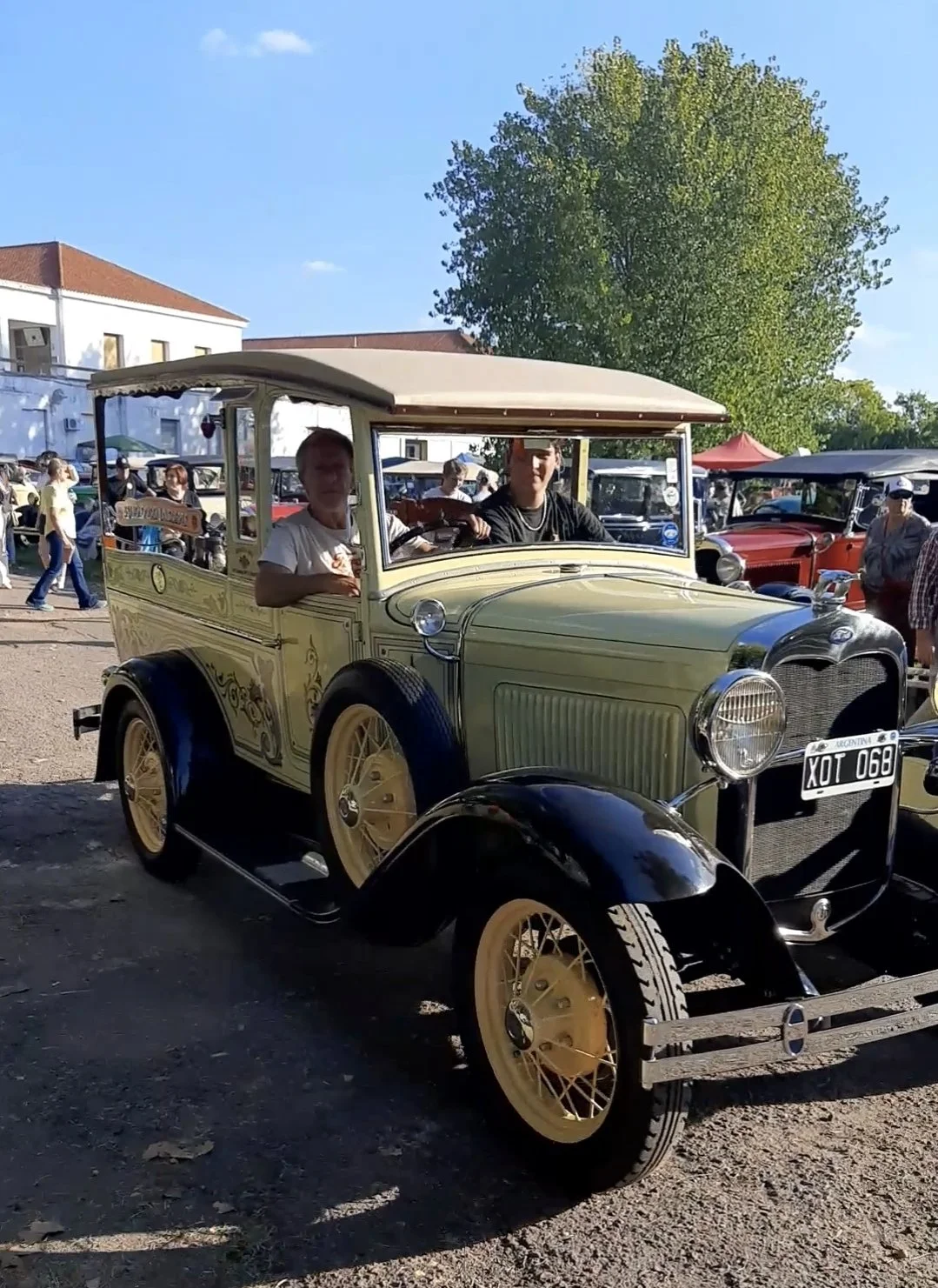 Auto antiguo en un evento con personas alrededor y un árbol grande de fondo.