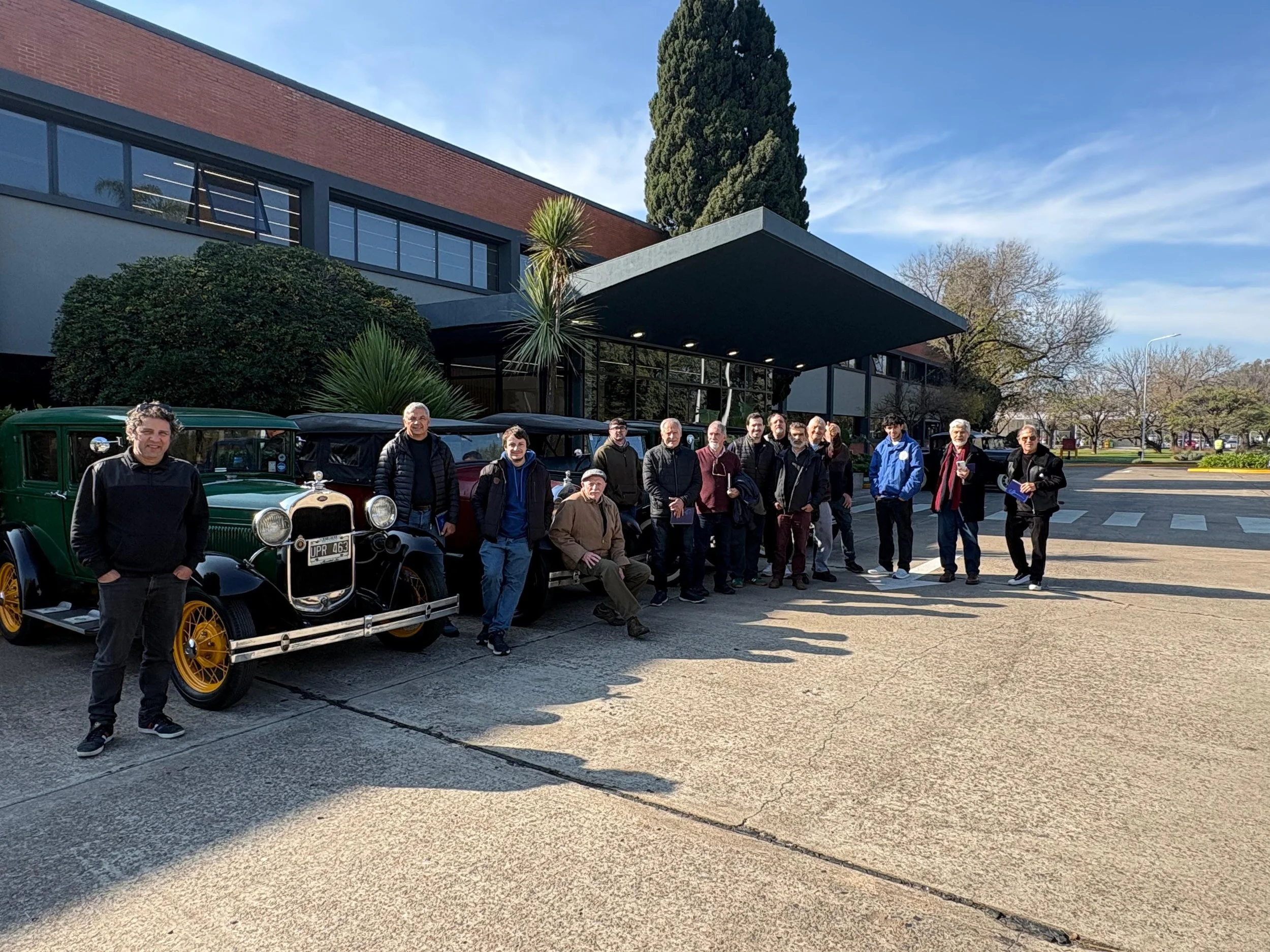 Grupo de personas posando frente a varios autos antiguos en un área al aire libre con árboles y un edificio moderno de fondo.