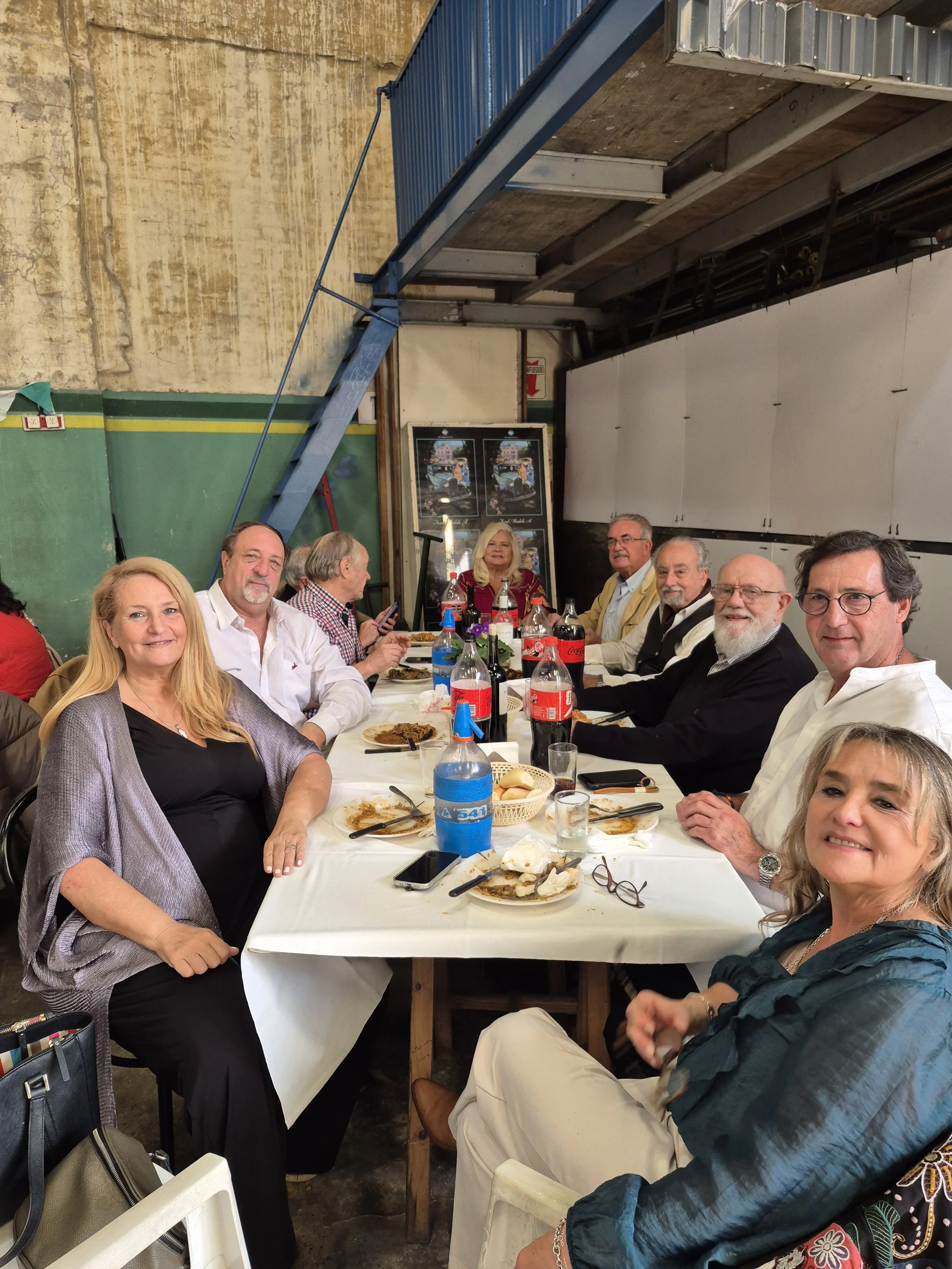 Grupo de personas sentadas alrededor de una mesa en un restaurante, algunos con platos de comida y bebidas, en un ambiente informal y ameno.