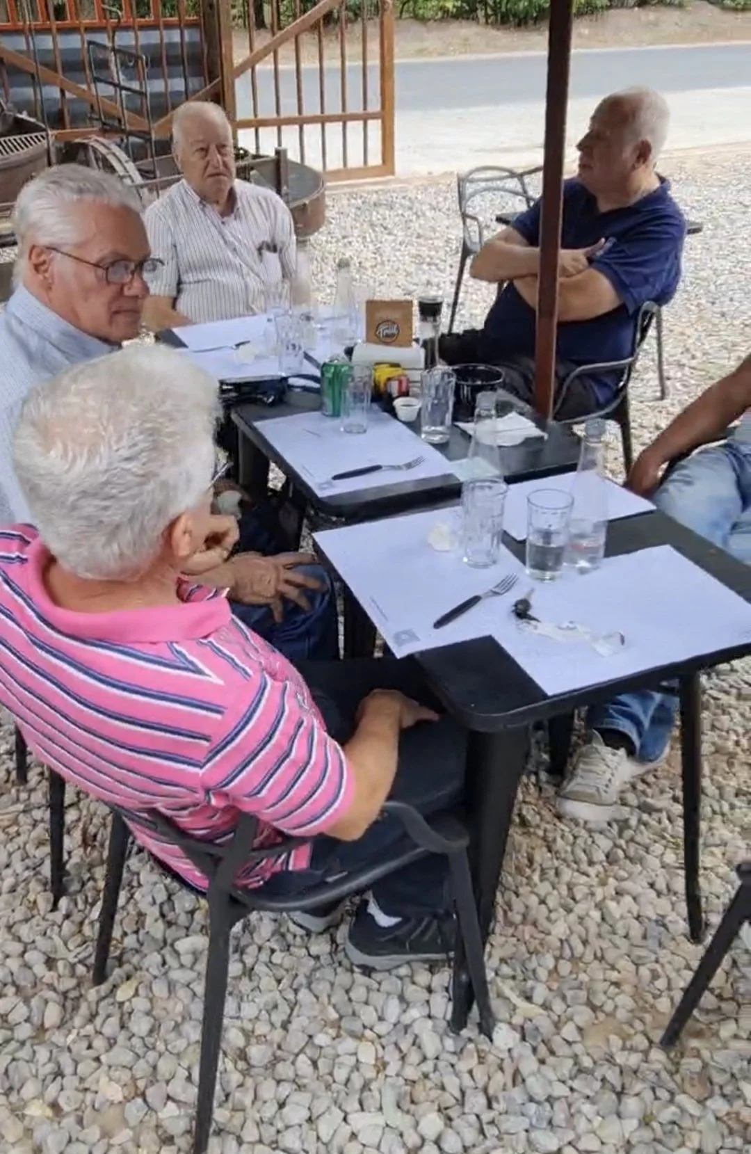 Grupo de personas mayores sentadas en una mesa al aire libre en un restaurante, con vasos y platos en la mesa, en un ambiente informal.