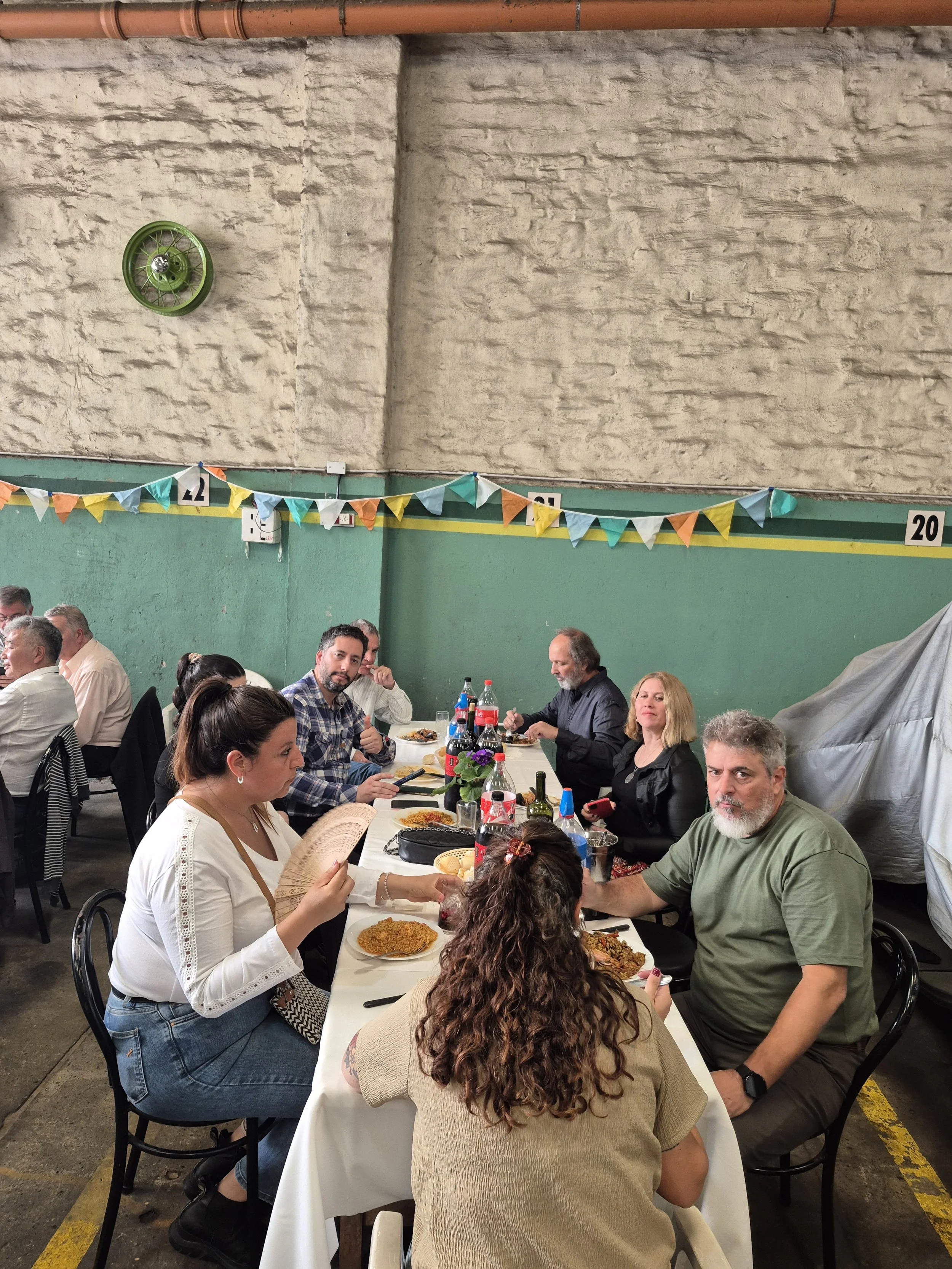 Personas sentadas en una mesa en un evento social, comiendo comida típica, en un espacio con paredes de piedra y decoraciones con banderines de colores.