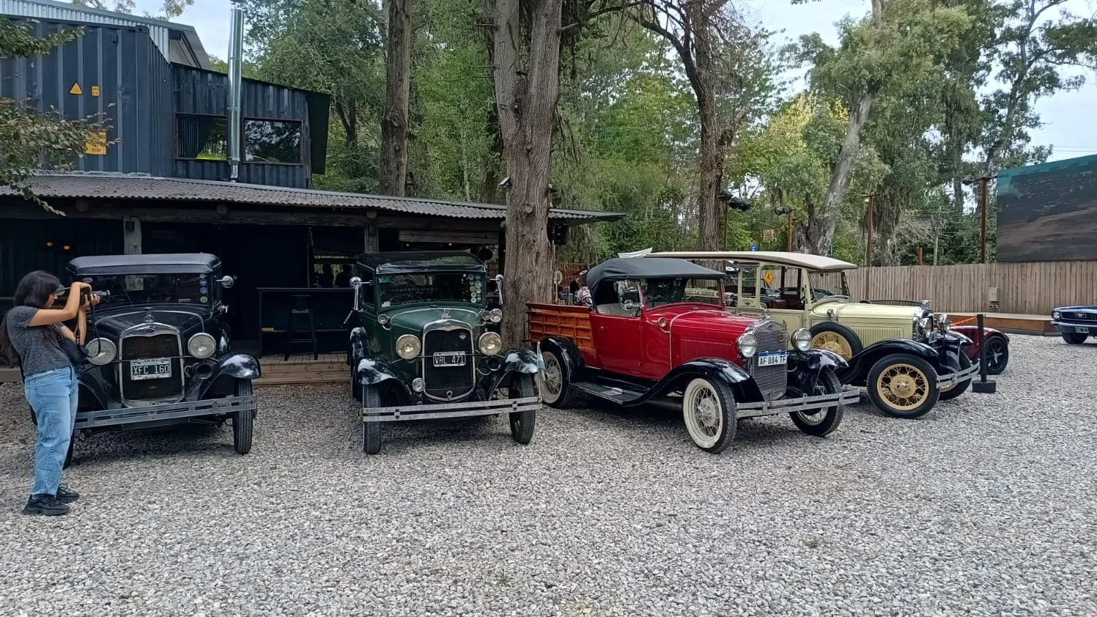 Colección de autos antiguos en exhibición, con una mujer tomando fotos y un fondo de árboles y estructura de madera.