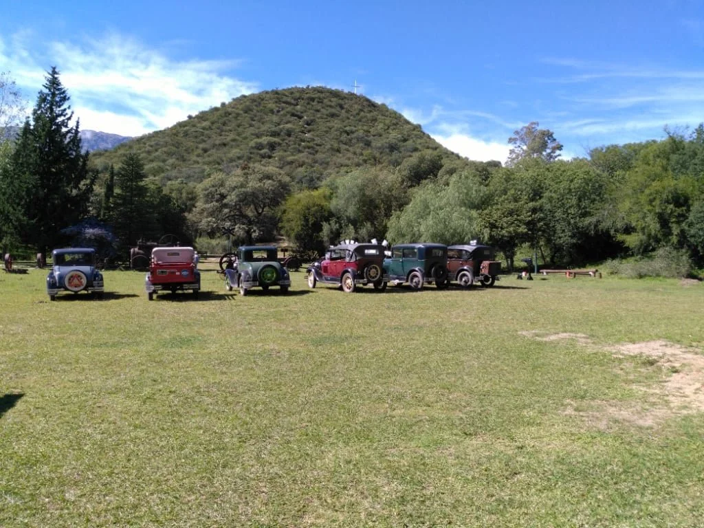 Varios autos clásicos estacionados en un campo rodeado de árboles y colinas bajo un cielo azul con algunas nubes.
