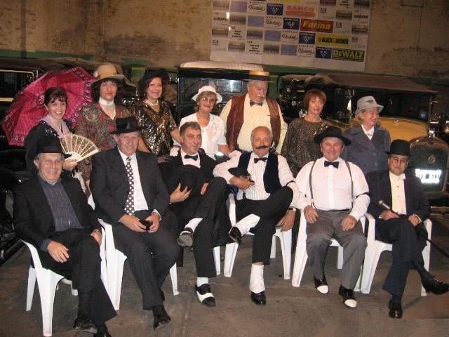 Grupo de personas vestidas con ropa de época, posando frente a autos antiguos en un ambiente que parece un evento temático vintage.