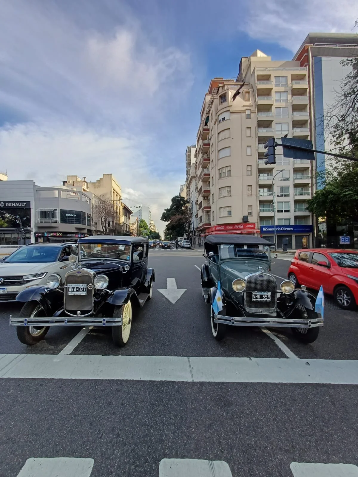 Calzada en una ciudad con autos modernos y dos autos antiguos clásicos en primer plano, edificios altos y tiendas al fondo, cielo parcialmente nublado.