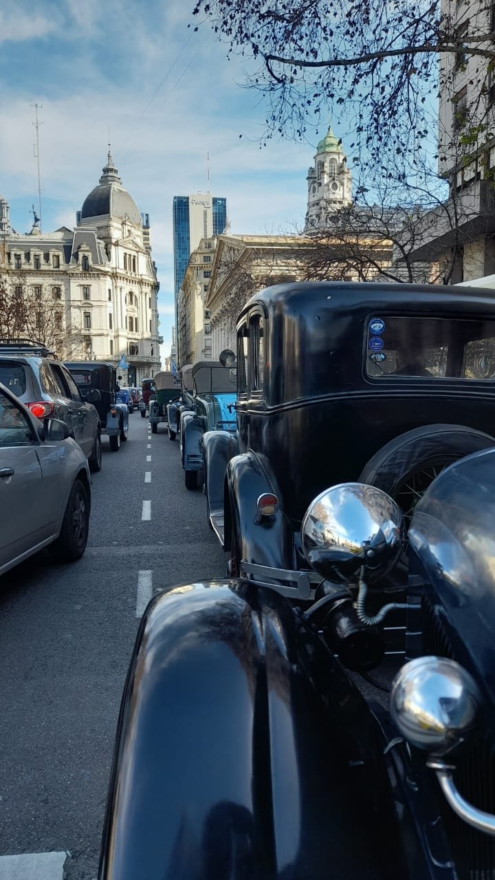 Vista de una calle en el centro de una ciudad, con autos antiguos en fila, algunos modernos, y edificios históricos y modernos de fondo, bajo un cielo parcialmente nublado.