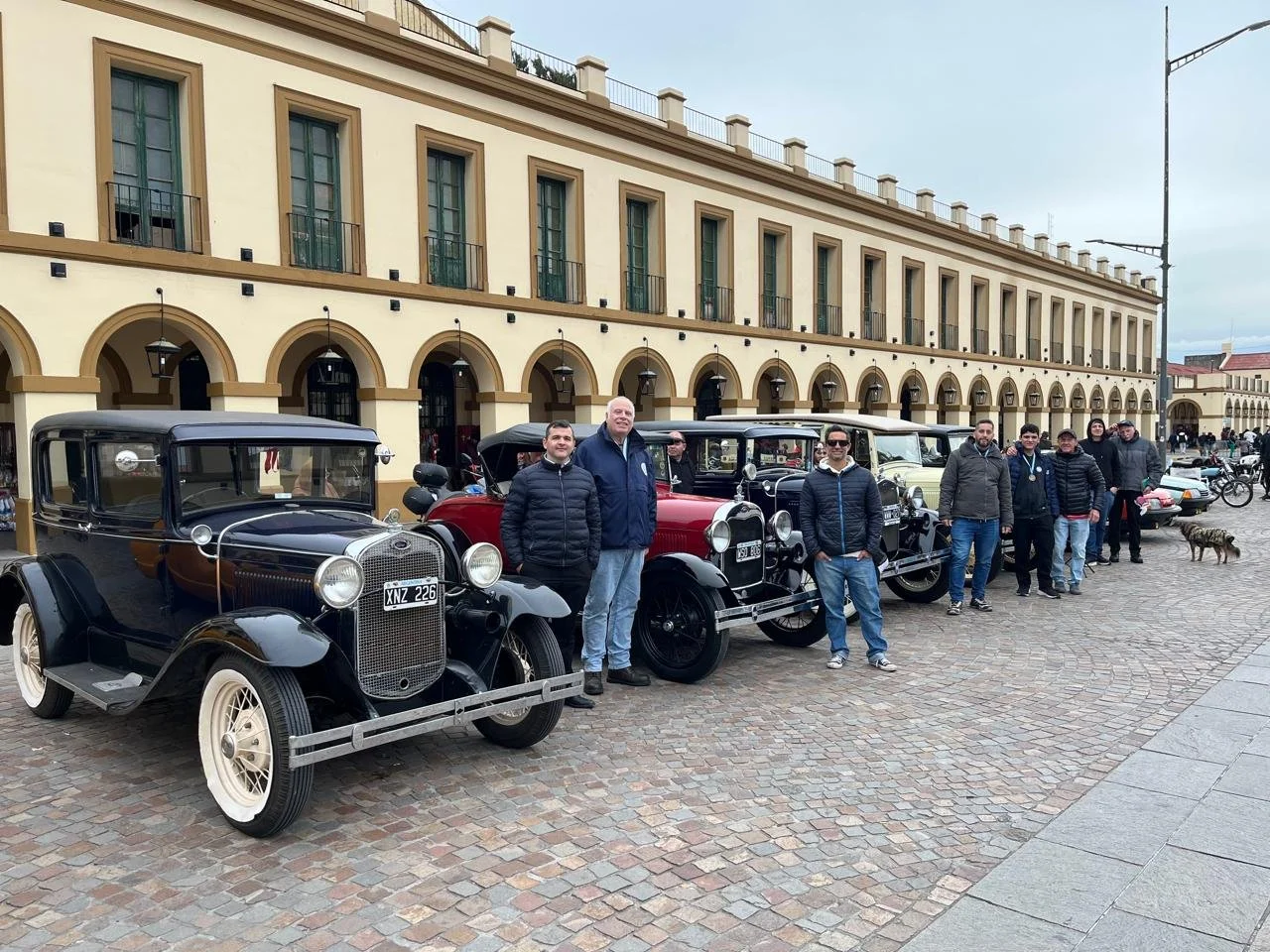 Grupo de personas posando junto a autos antiguos en una plaza en día nublado.
