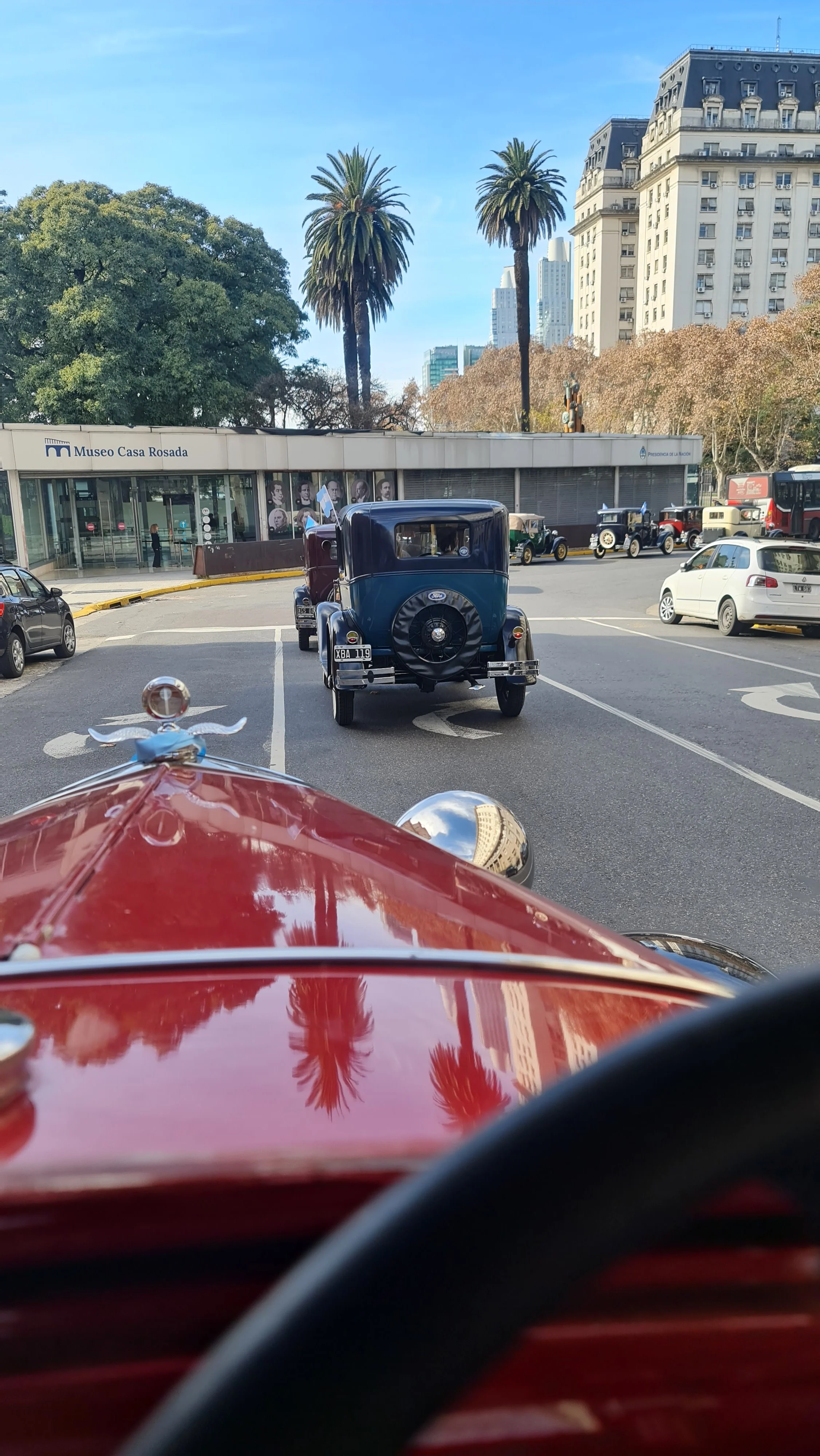 Vista desde el interior de un automóvil rojo clásico, mostrando el tablero y el espejo retrovisor. Delante hay otro coche clásico de color negro y más autos en movimiento en una ciudad con edificios altos y palmeras, y una cerca del Museo Casa Rosada