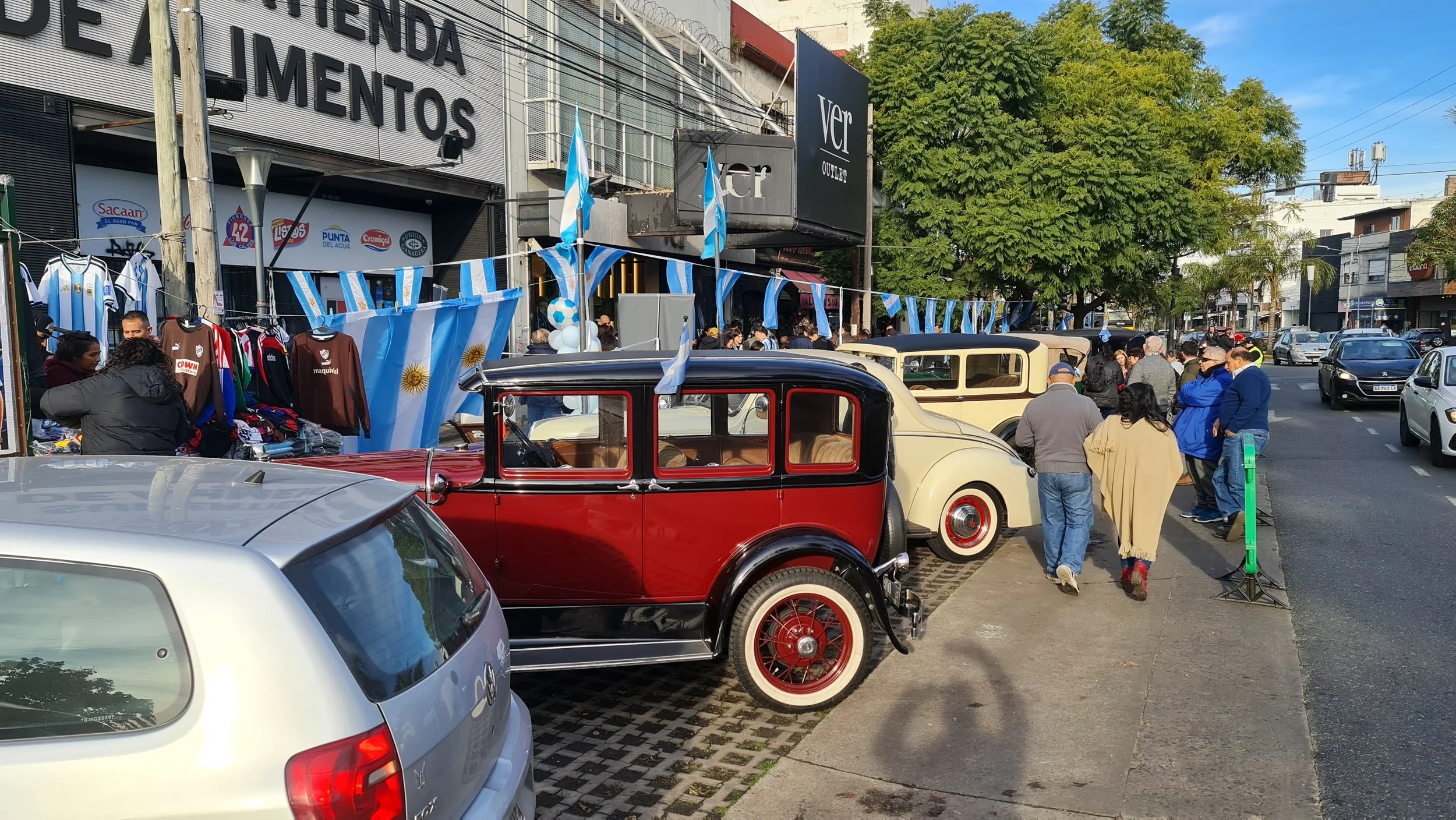 Celebración de la selección Argentina con banderas, globos y autos antiguos en la calle, con muchas personas disfrutando del evento y decoraciones en azul y blanco.