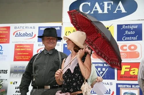 Una pareja vestida con ropa vintage, con sombreros y un paraguas, posando frente a un cartel publicitario.