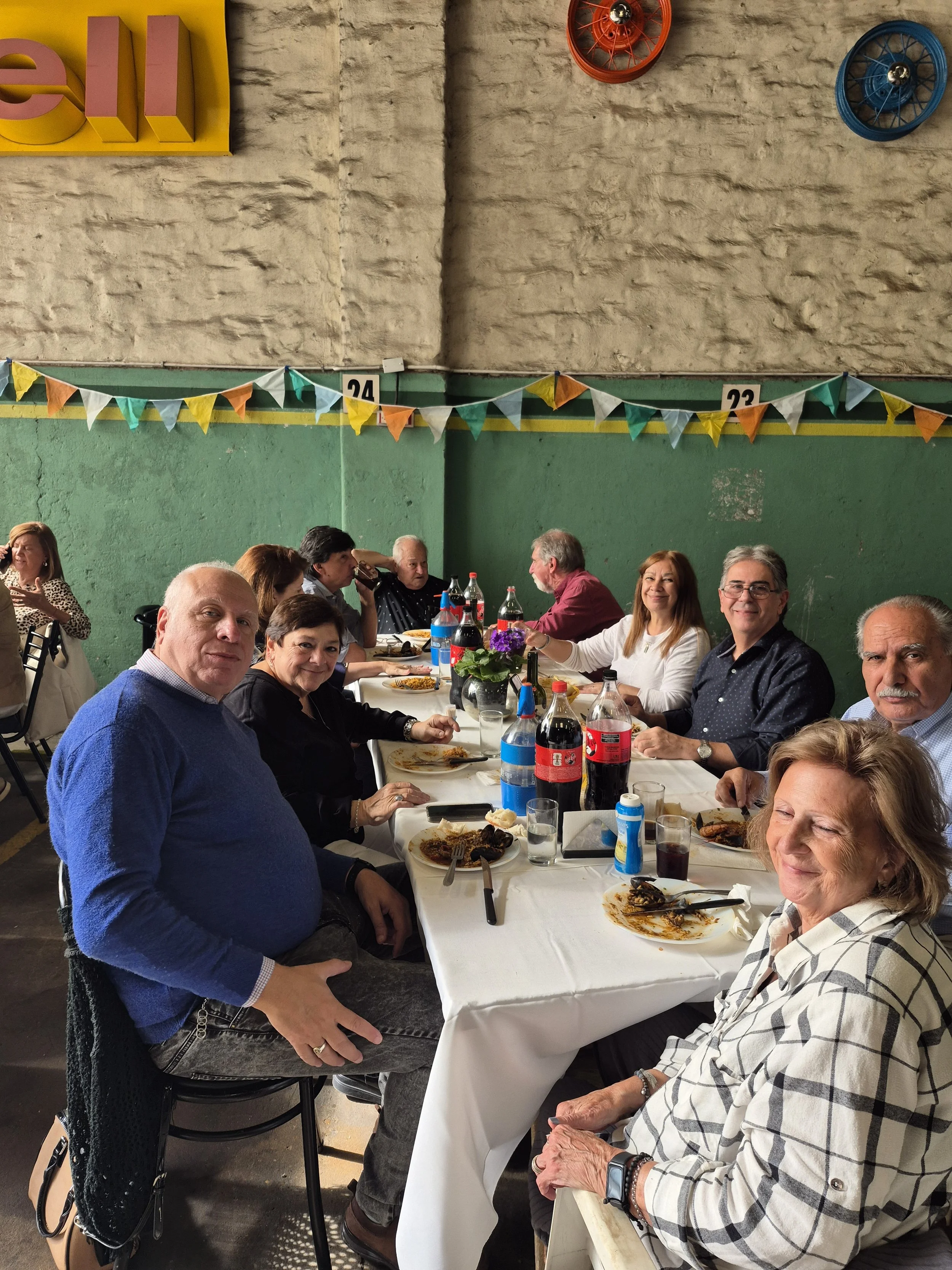 Personas reunidas en una mesa comiendo y socializando en un ambiente festivo con decoración de banderines y relojes de pared de colores vivos.