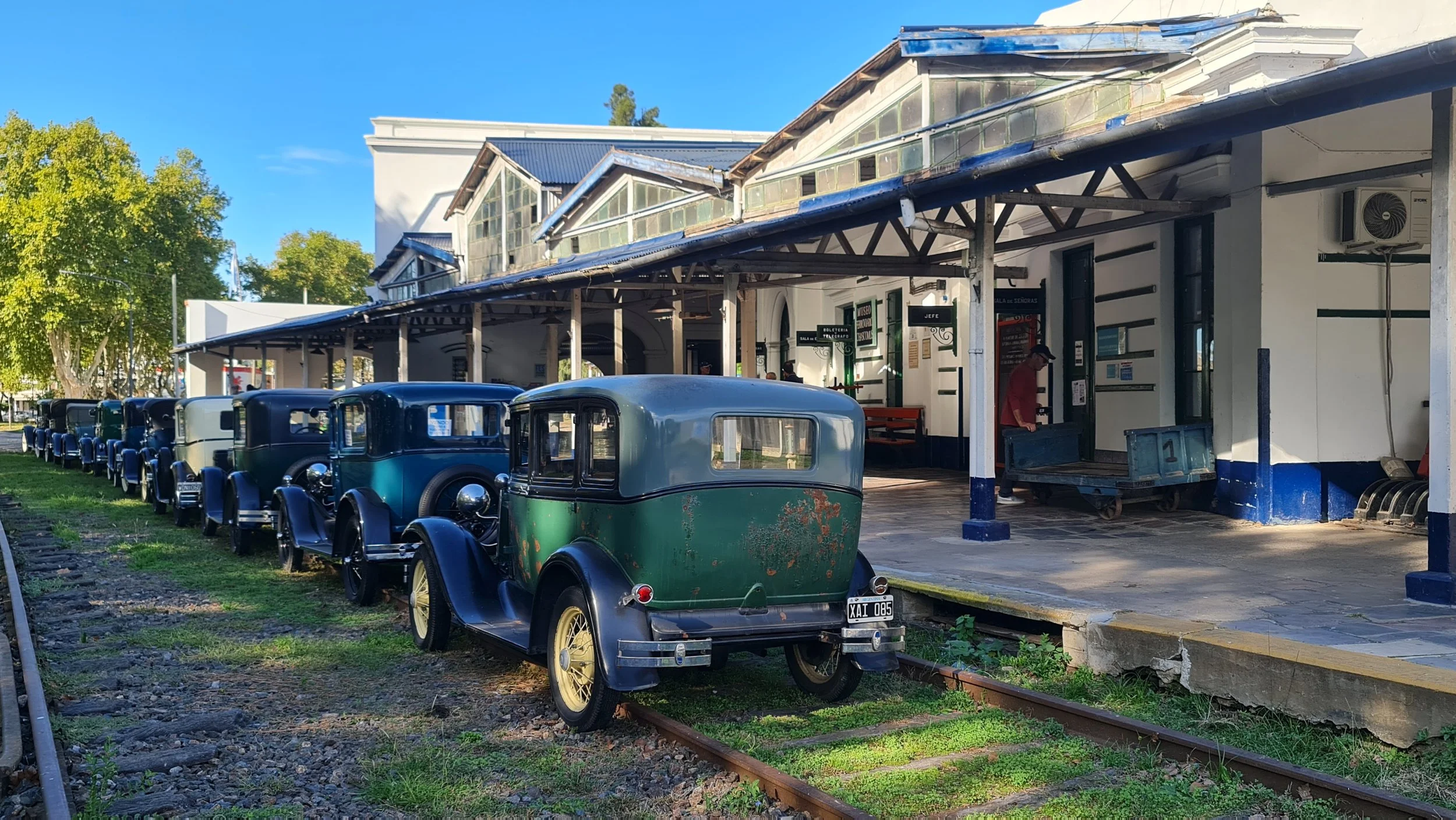 Estación de tren antigua con coches de época en la vía y un edificio con vestíbulo y taquillas.