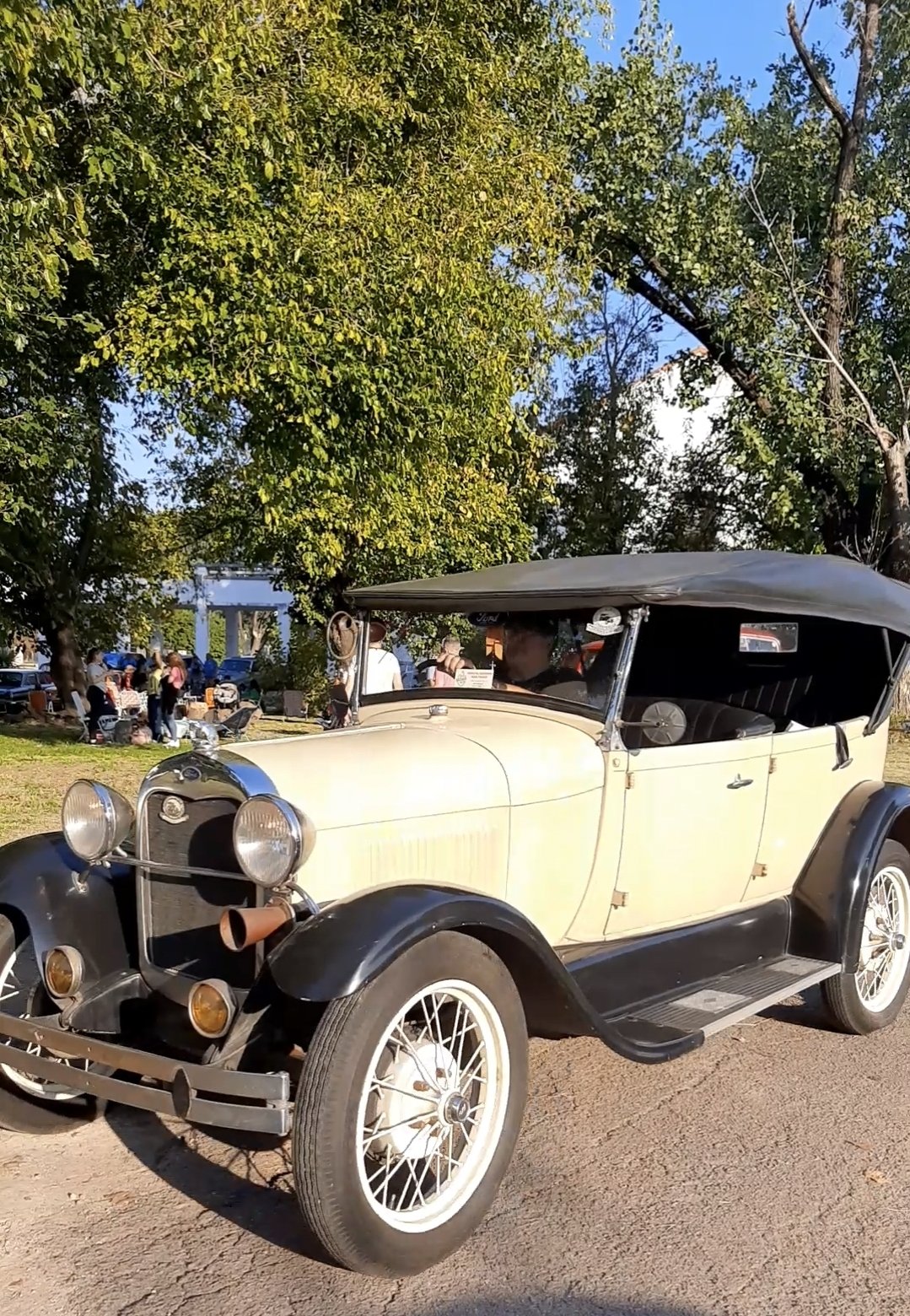 Coche clásico de color beige con detalles en negro y techo de lona, estacionado en un parque con árboles y personas en el fondo.