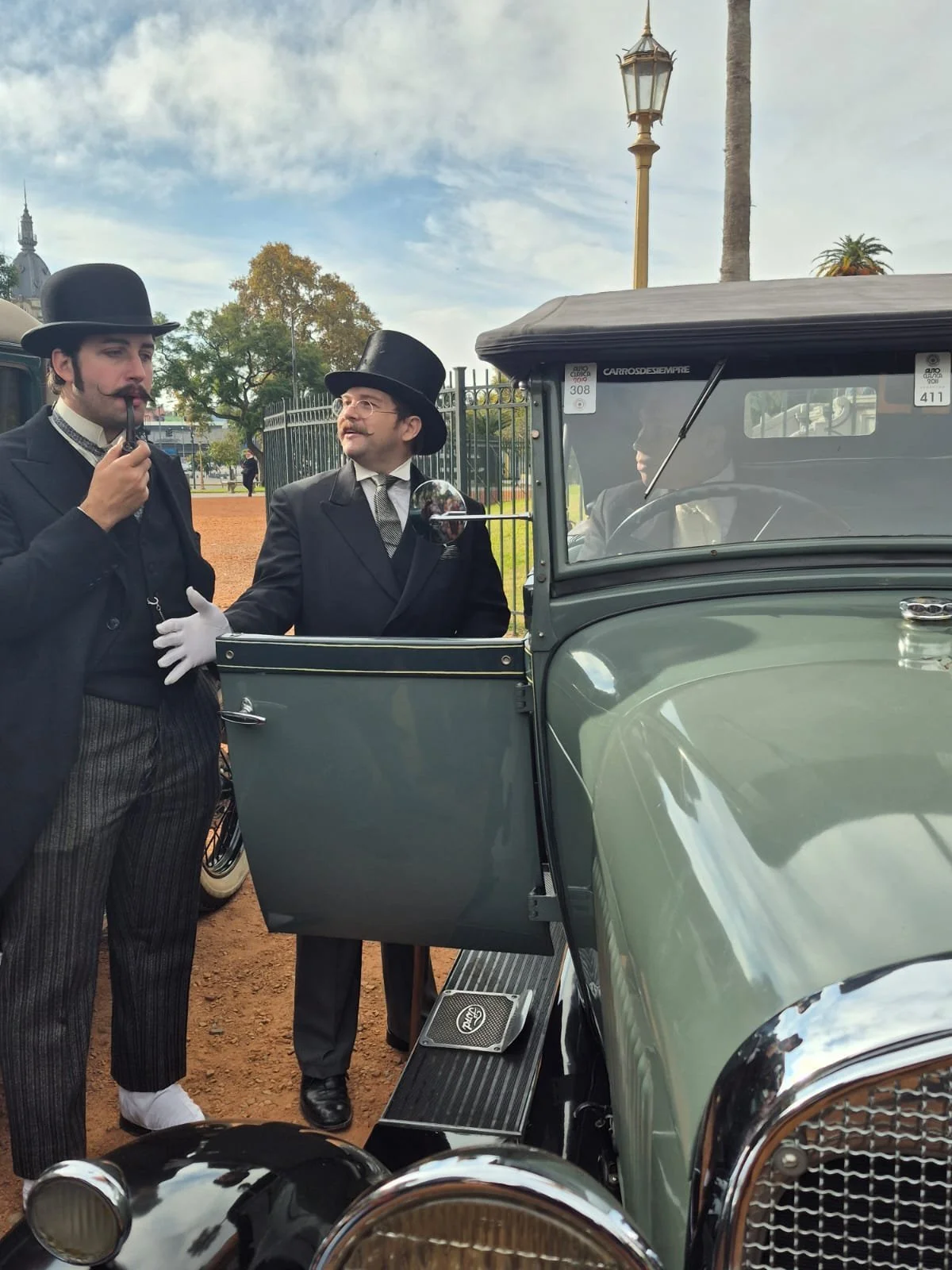 Dos hombres con trajes antiguos y sombreros charlando junto a un coche clásico.