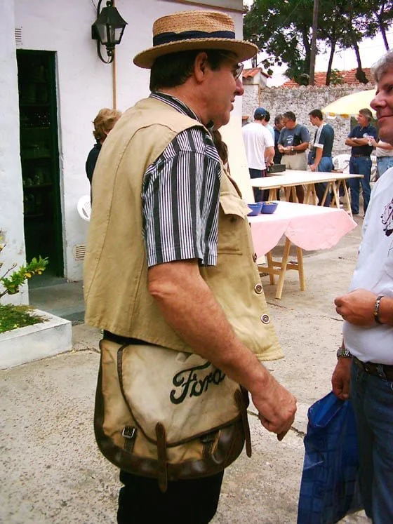 Un hombre con sombrero, camiseta a rayas, y chaleco, lleva una bolsa de cuero con logo Ford, participa en un evento al aire libre con varias personas en el fondo.