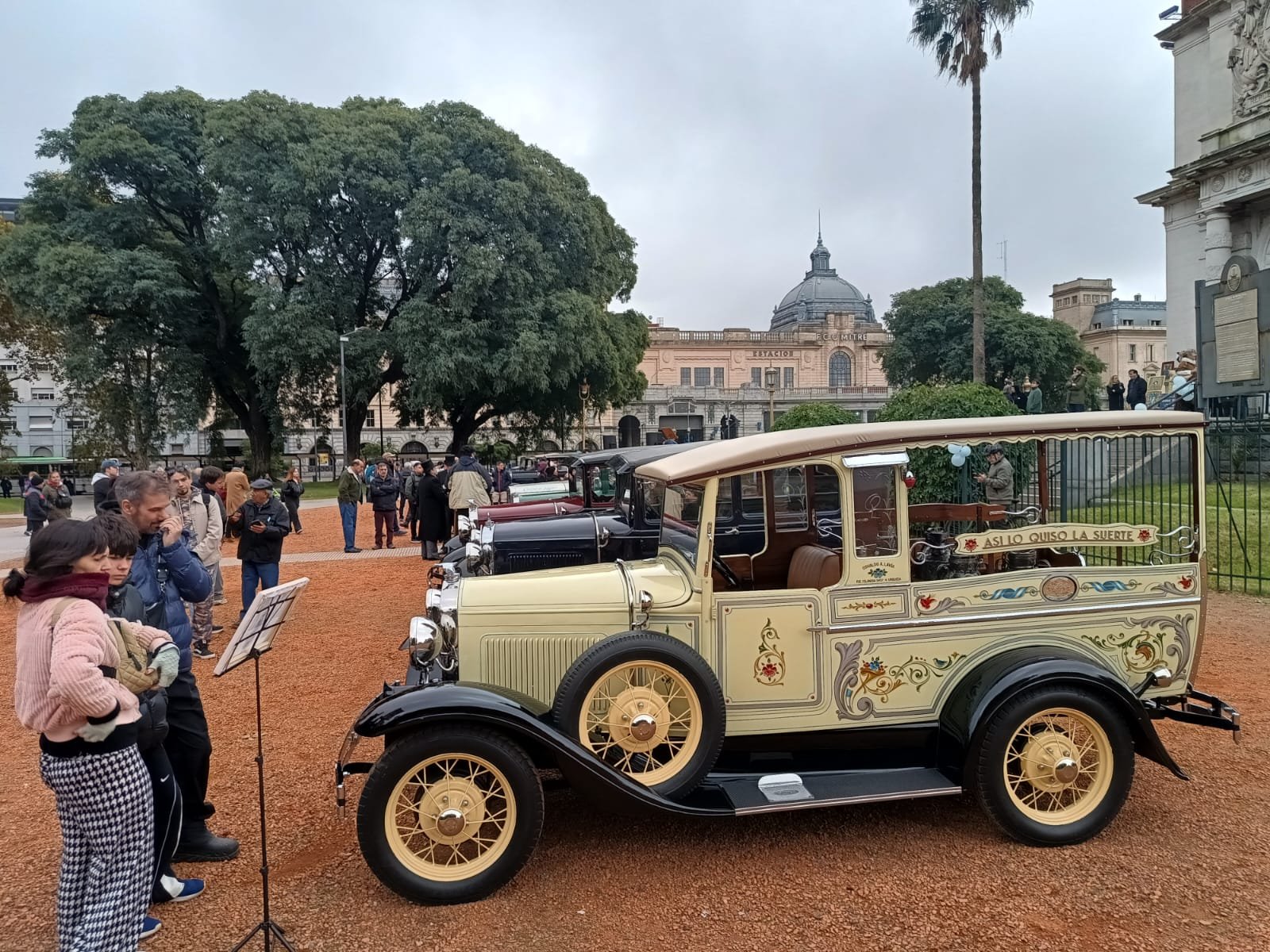 Autos antiguos en una exposición en un parque con personas observando y en el fondo árboles, un edificio y un cielo nublado.