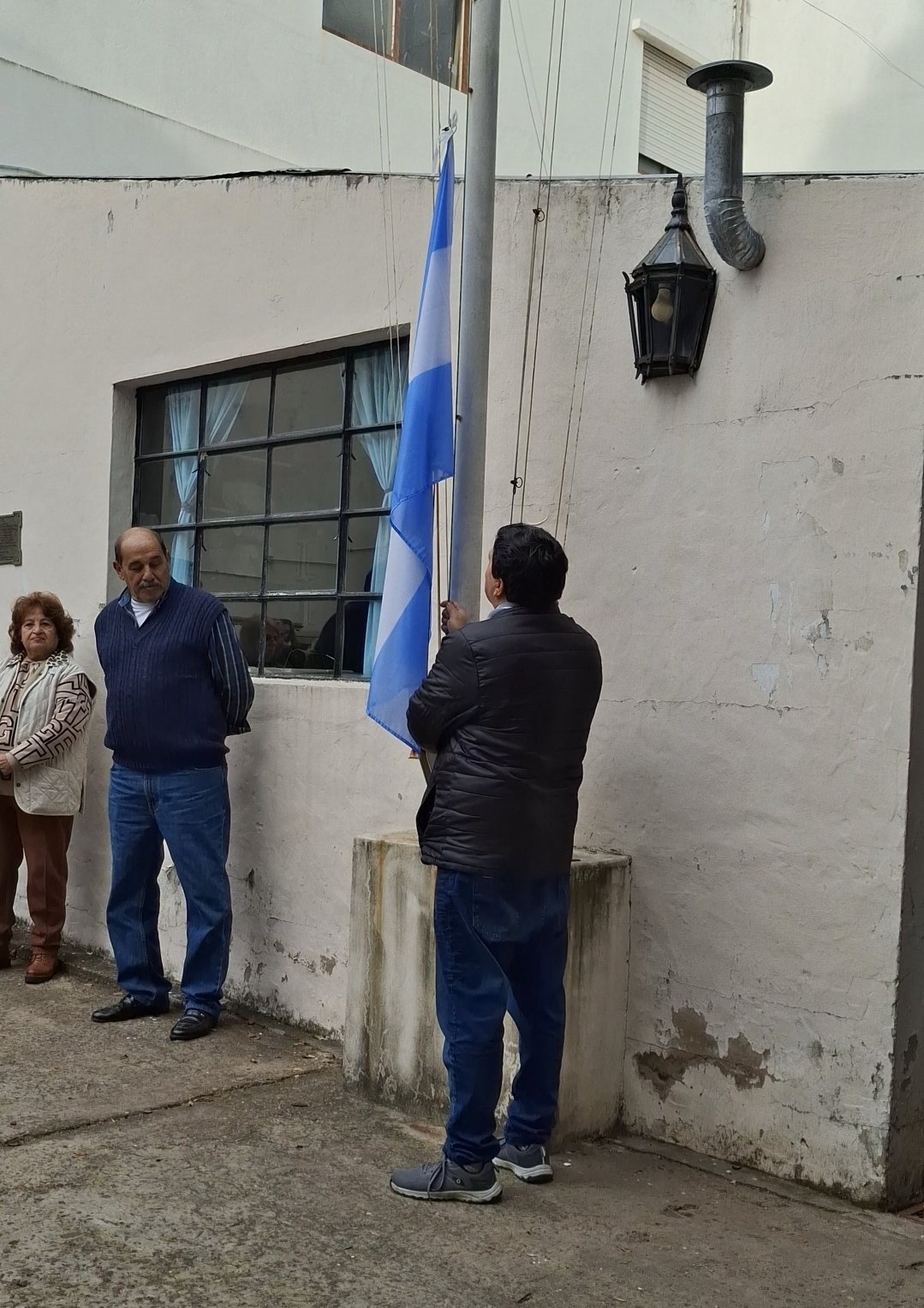 Hombre colocando la bandera de Argentina en un asta, acompañado por dos personas observando la ceremonia en el exterior de un edificio.