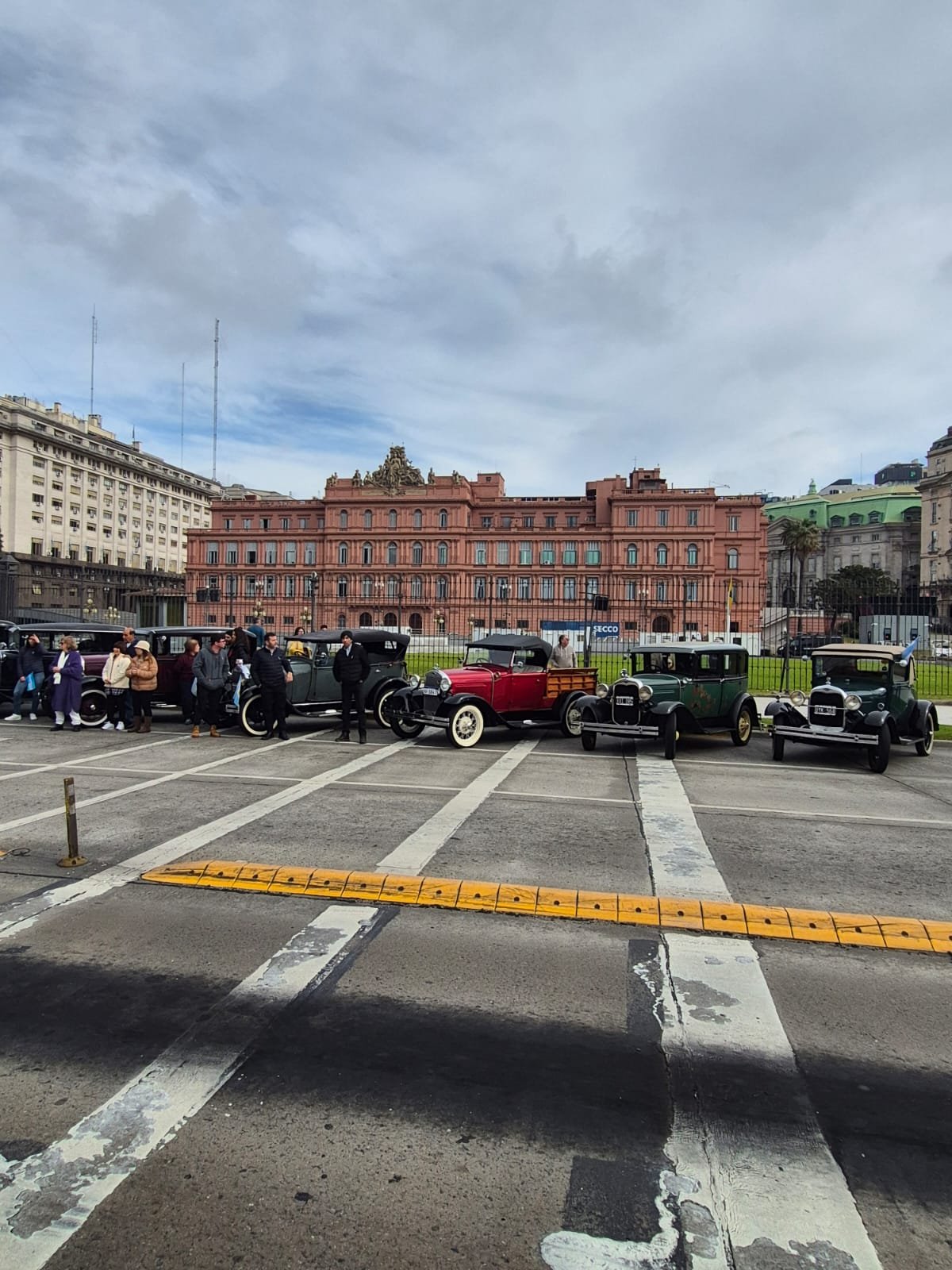 Autos antiguos y personas en un parque en una ciudad con edificio rosa y cielos nublados.