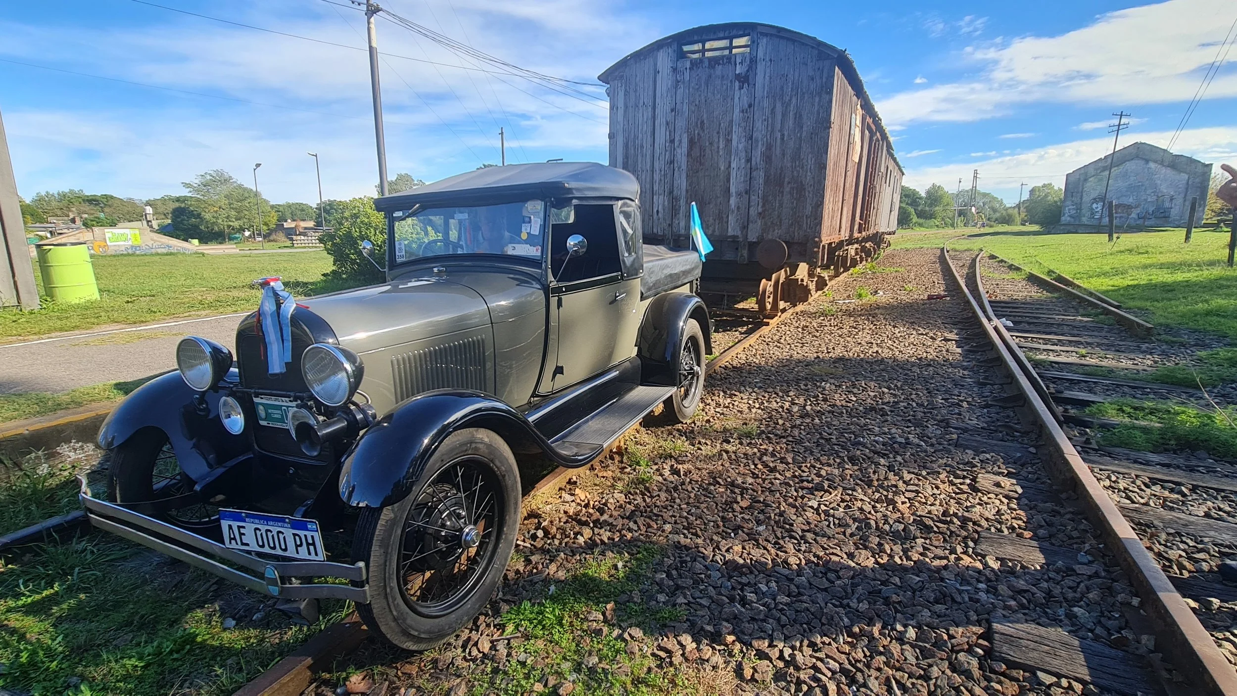 Un coche antiguo negro con techo blando en la orilla de las vías del tren, junto a un vagón de tren de madera envejecido, en un entorno rural con campos verdes y edificios a lo lejos bajo un cielo azul con nubes.
