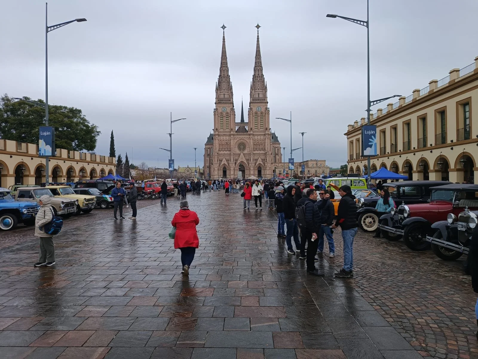 Plaza frente a la catedral en Luján con coches clásicos en exhibición y personas caminando en un día nublado.