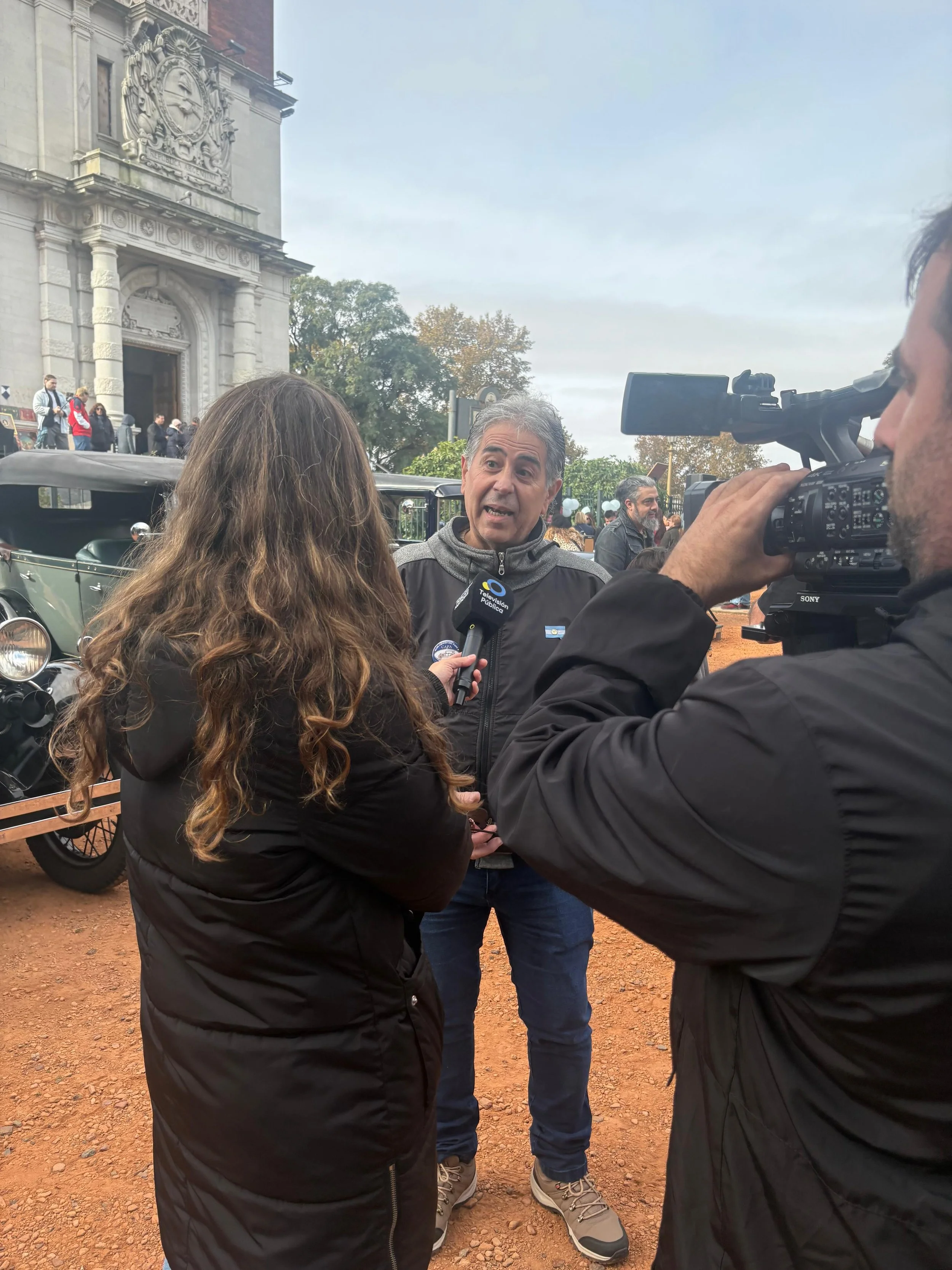 Hombre siendo entrevistado por periodistas en una plaza con edificio antiguo y árboles en el fondo.