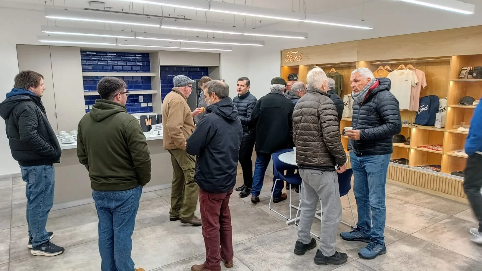Grupo de personas conversando en un espacio interior, con exhibiciones de souvenirs y merchandise de Ford en el fondo, algunos tomando café.