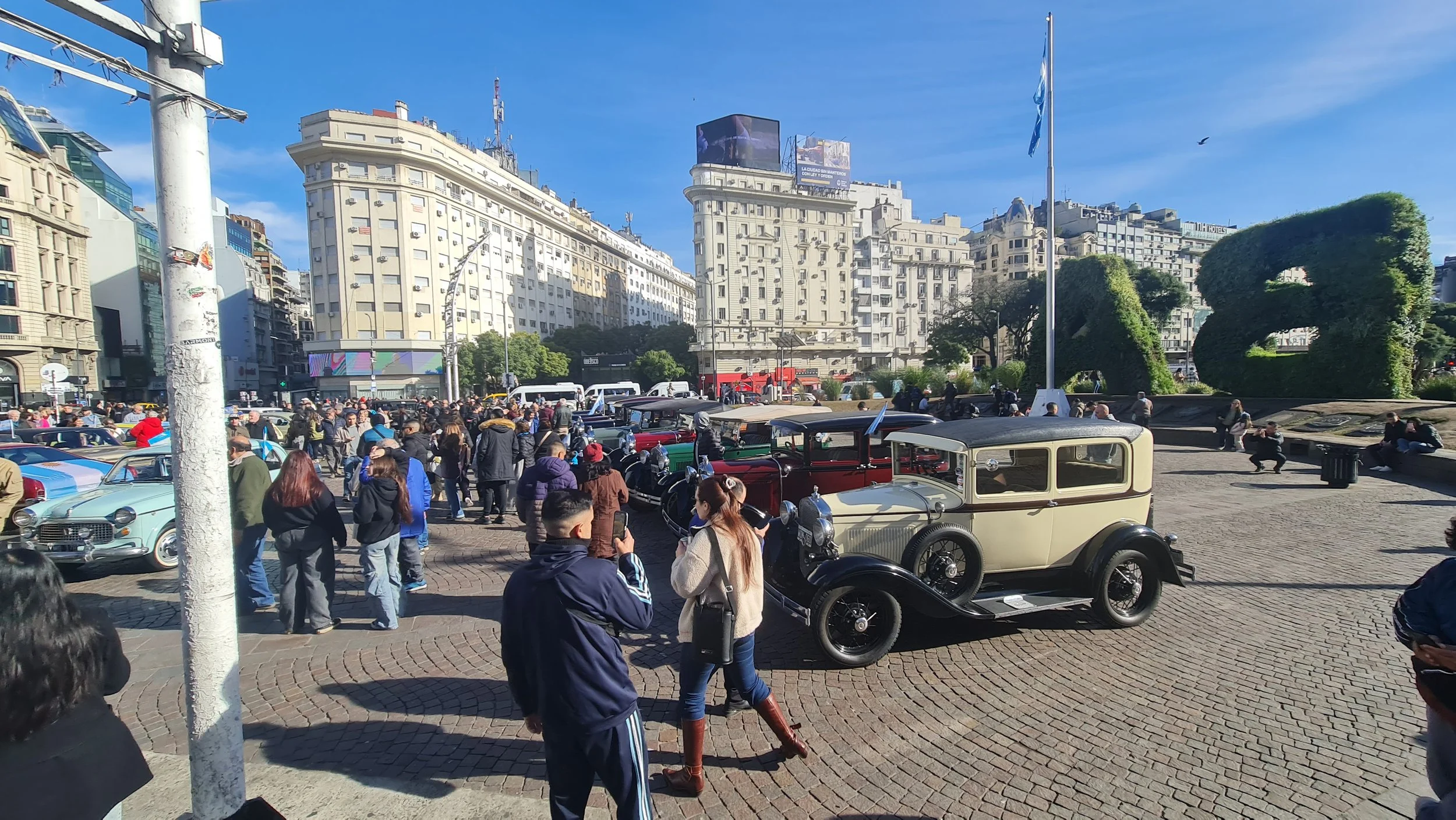 Una plaza concurrida con autos antiguos en exhibición y muchas personas paseando. Edificios altos y un cielo despejado en el fondo.