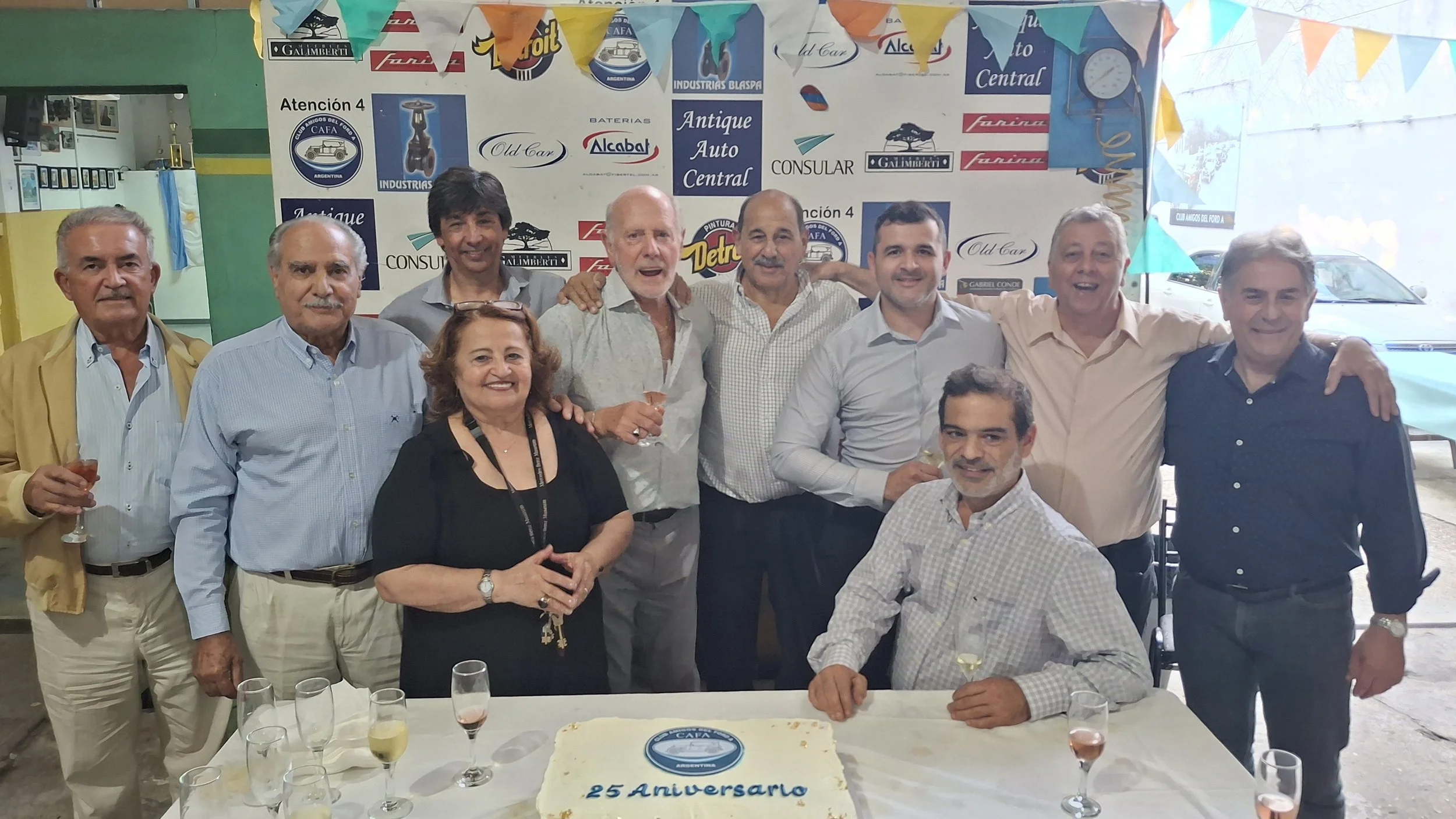 Un grupo de personas celebrando un 25 aniversario, con una mesa decorada y un pastel en primer plano, en un lugar decorado con banners y logos de autos antiguos.