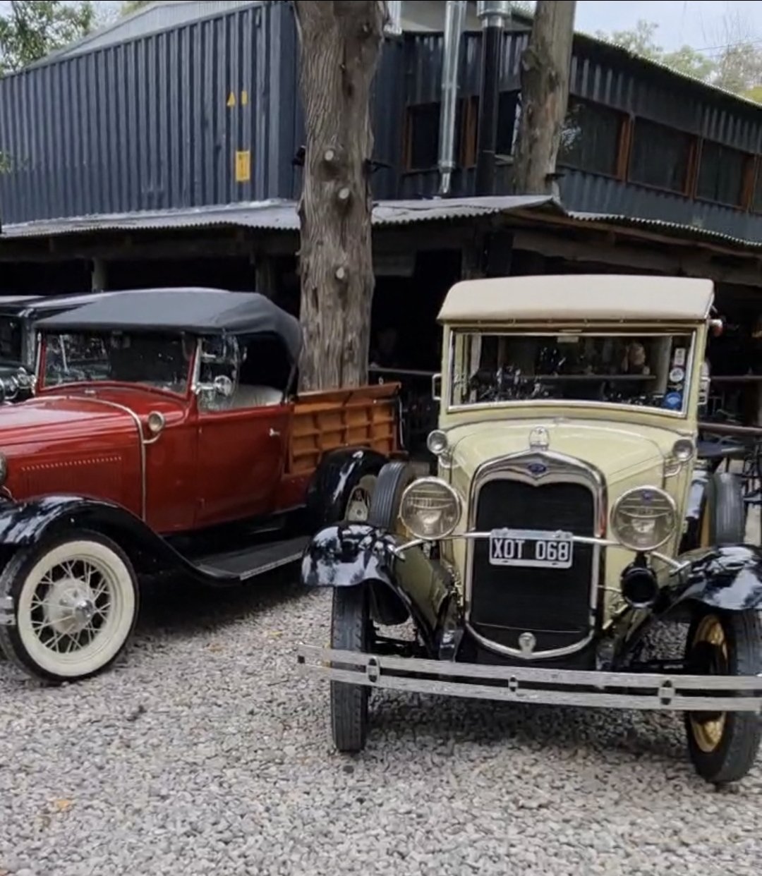 Duatros autos clásicos estacionados en un lugar con árboles y estructura de madera y metal en el fondo. Uno es rojo con capota negra y el otro es amarillo con una parrilla delantera antigua.