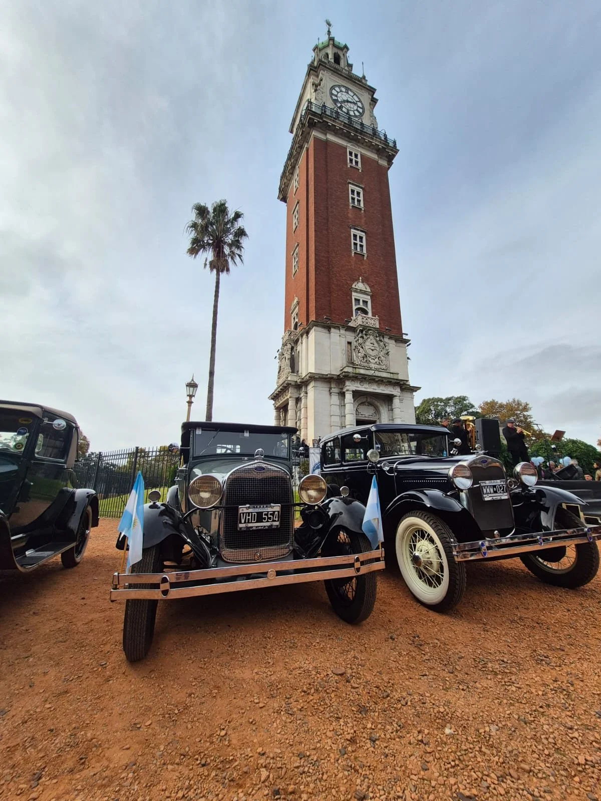 Carros antiguos en un evento frente a una torre con reloj, en un día nublado.