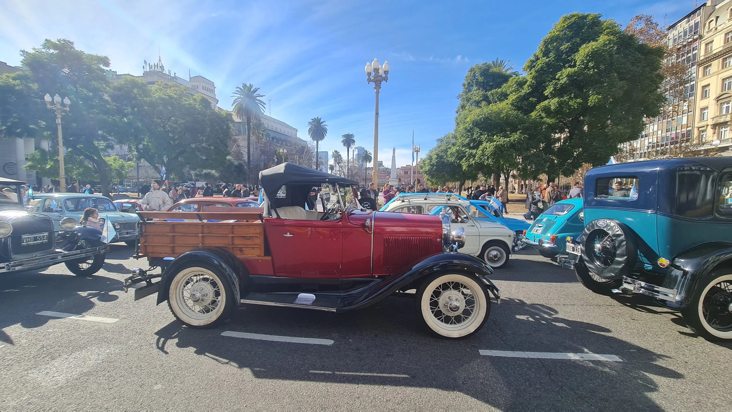 Autos antiguos en una plaza pública con árboles y gente, en un día soleado con un cielo azul.