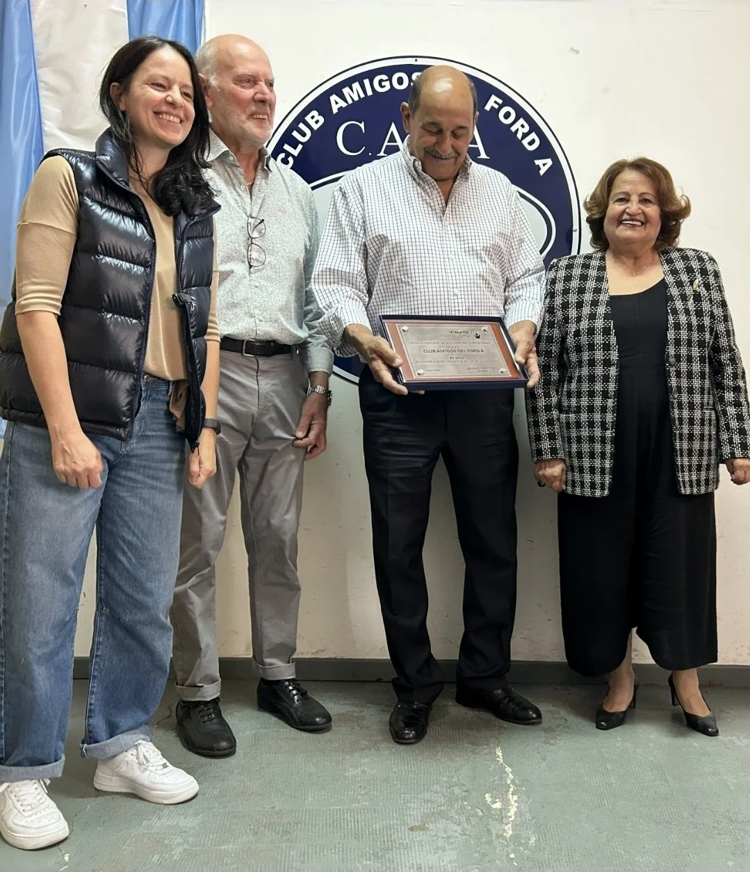 Cinco personas en una ceremonia, una de ellas presenta un certificado a otra, fondo con logotipo del club de automovilismo 'Club Amigos Ford A'.