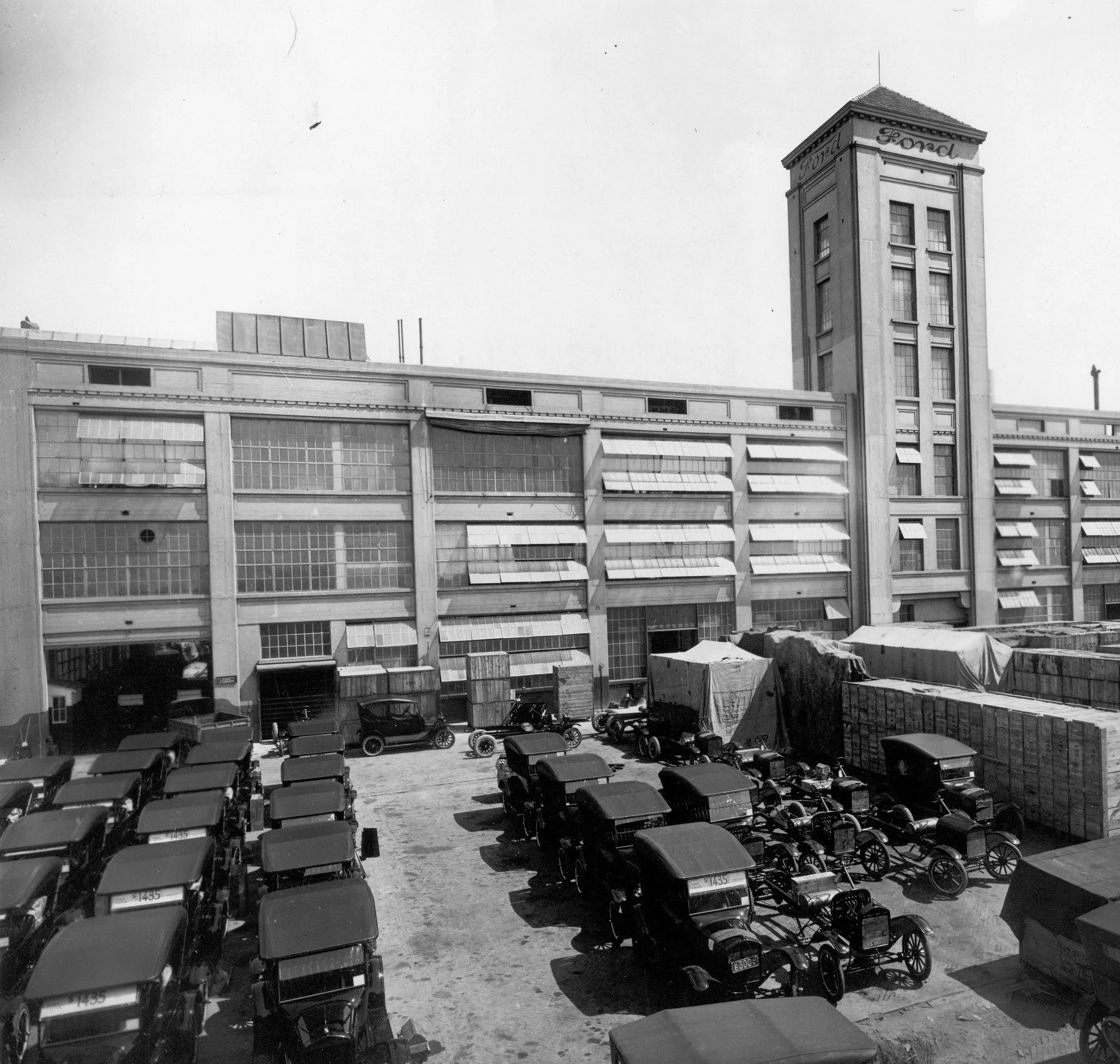 Edificio de varios pisos con fachada de ventanas, un letrero que dice Ford en la parte superior de una torre, y un estacionamiento con autos en la parte inferior. Se observan toldos y una estructura de lona en el área del estacionamiento.