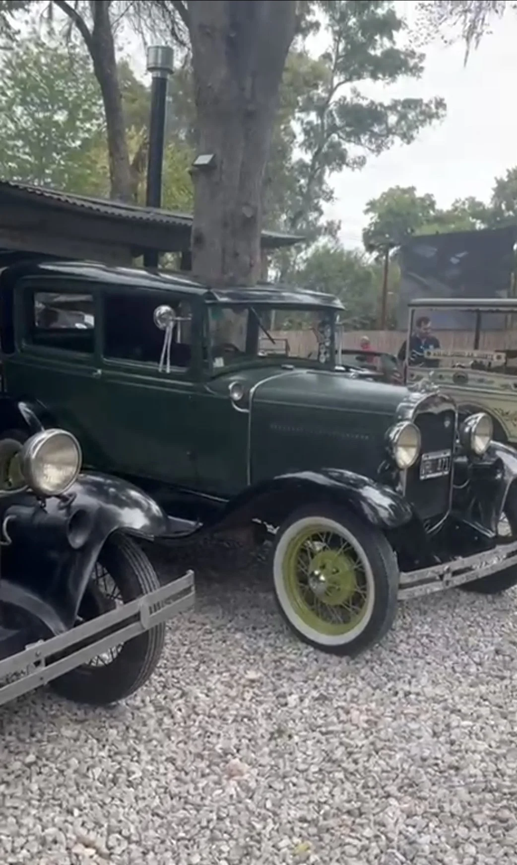 Automóviles antiguos, entre ellos un coche de color verde y otros de estilo vintage, exhibidos al aire libre con árboles de fondo.