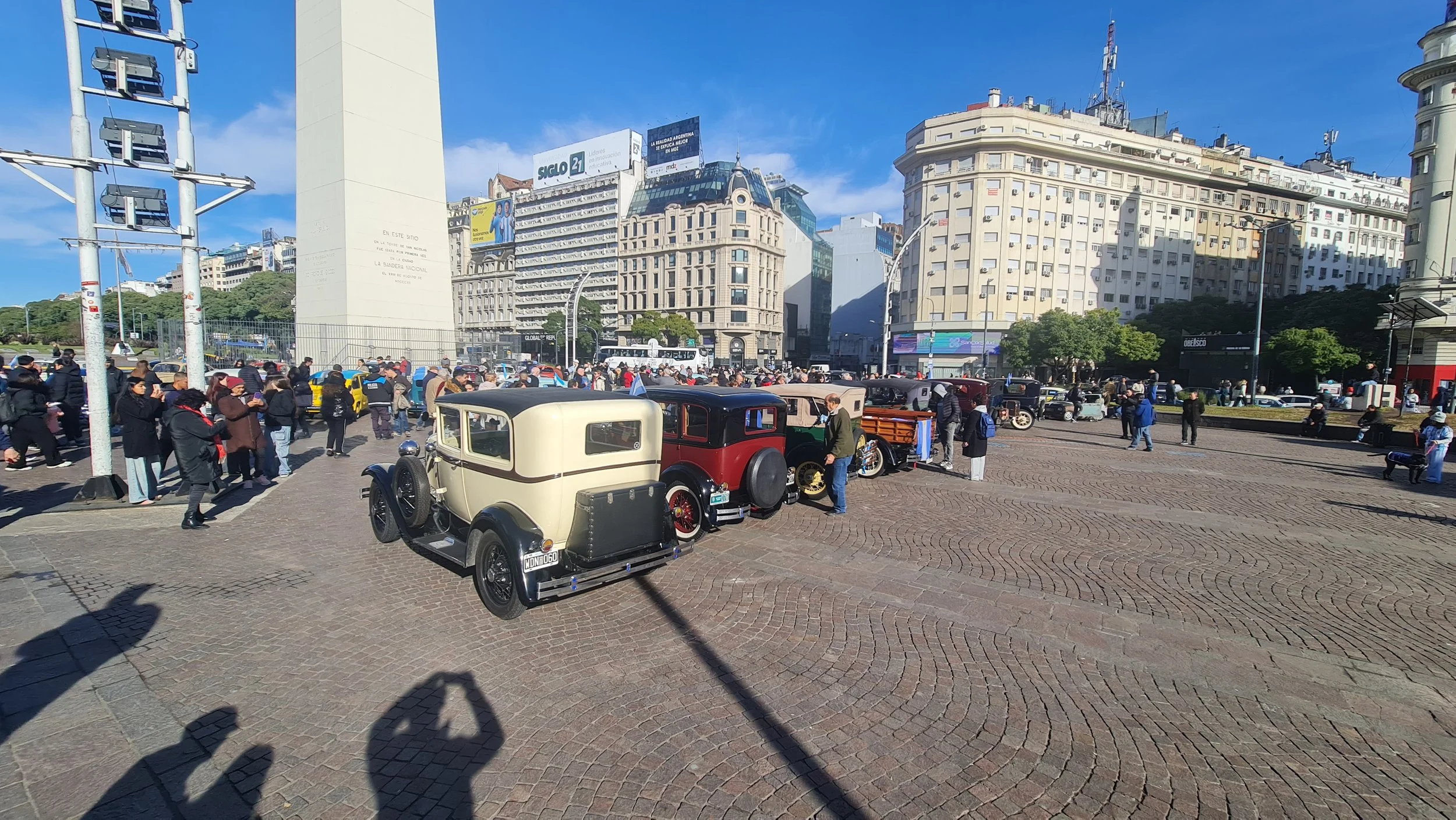 Plaza con coches antiguos y muchas personas en un evento público en una ciudad con edificios altos y un cielo despejado.