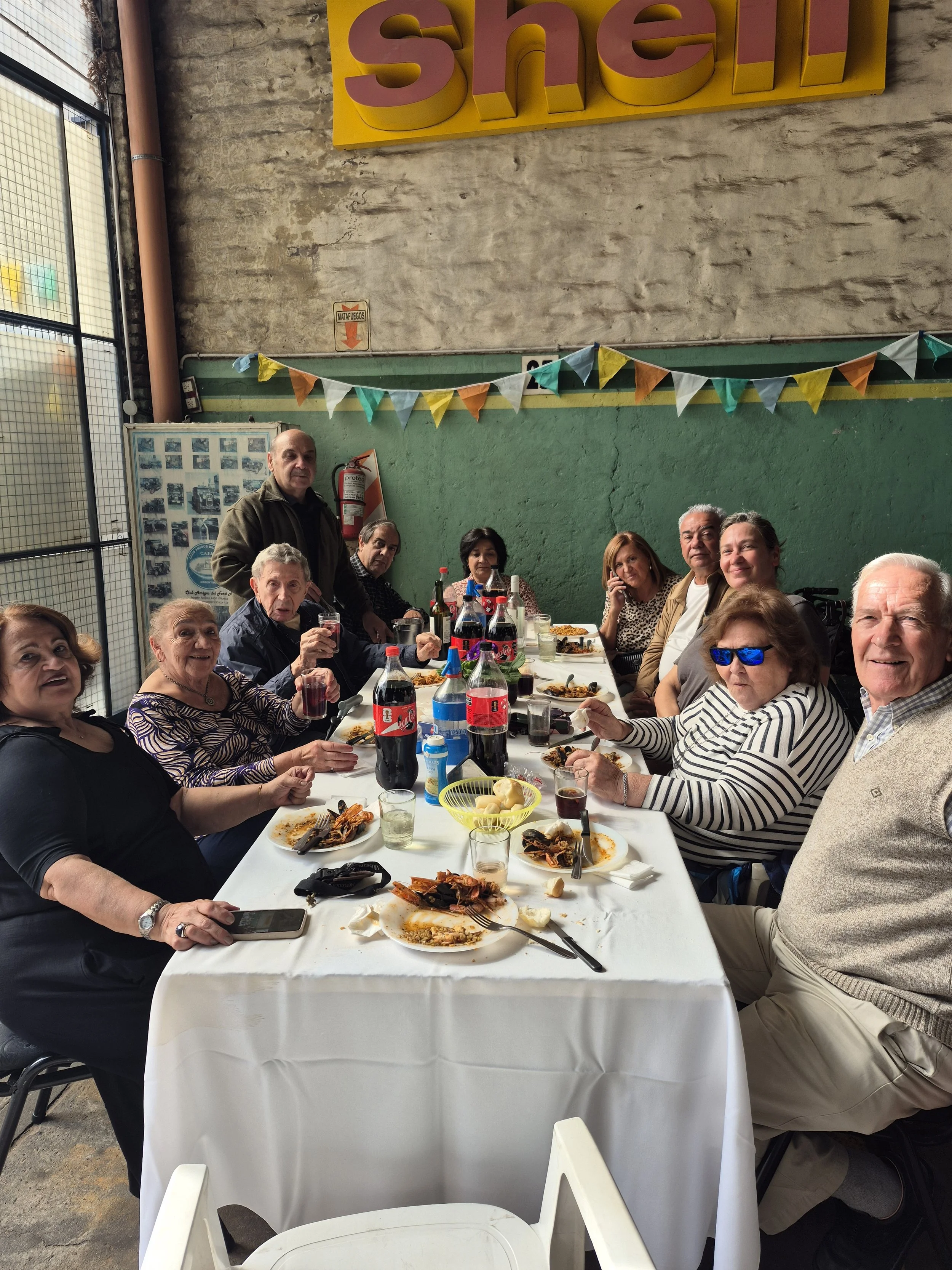 Grupo de personas sentadas en una mesa disfrutando una comida en un espacio cerrado con paredes de ladrillo y cartel amarillo con letras rosas que dice 'SHIEL'.