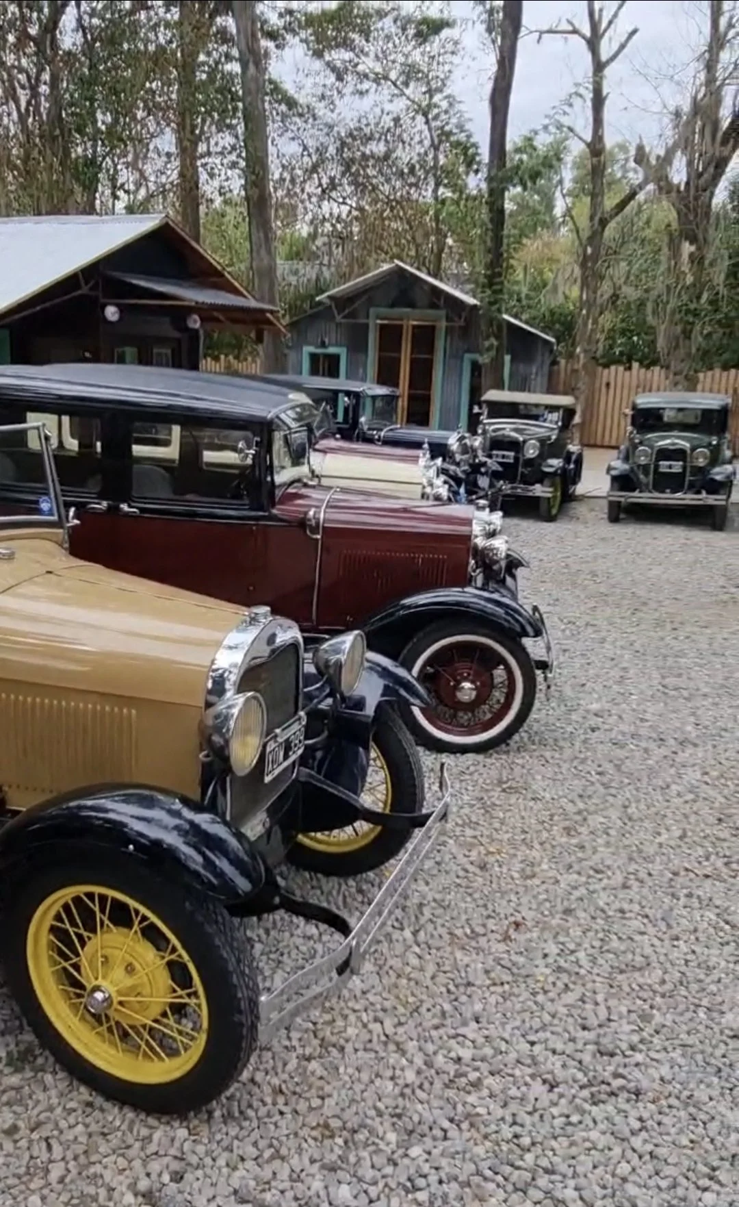 Autos antiguos estacionados en un espacio de grava, con casas de madera y árboles al fondo.