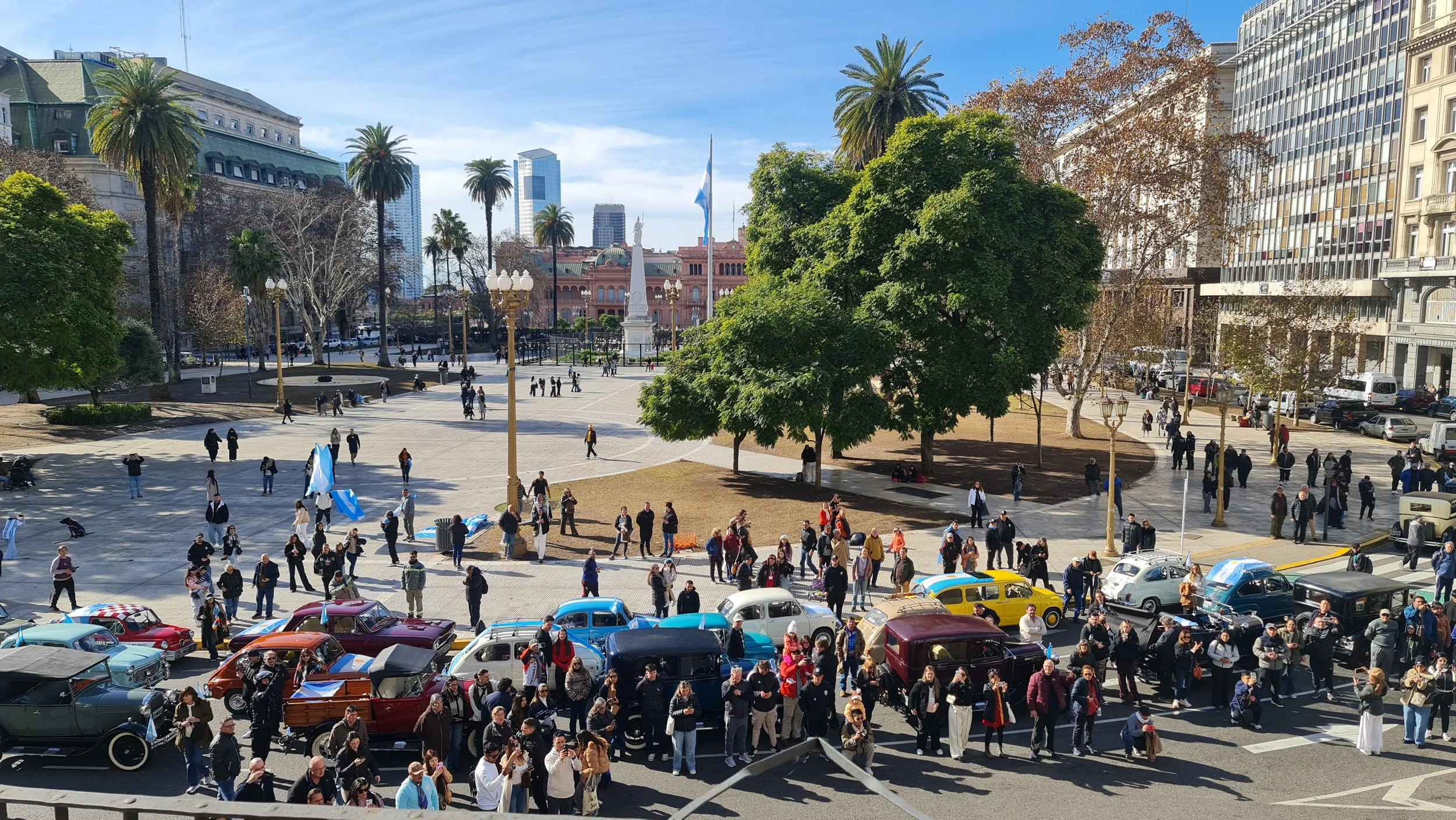 Vista de una plaza con muchas personas, autos antiguos y árboles, con edificios altos al fondo y un monumento en el centro onde ondea la bandera argentina.