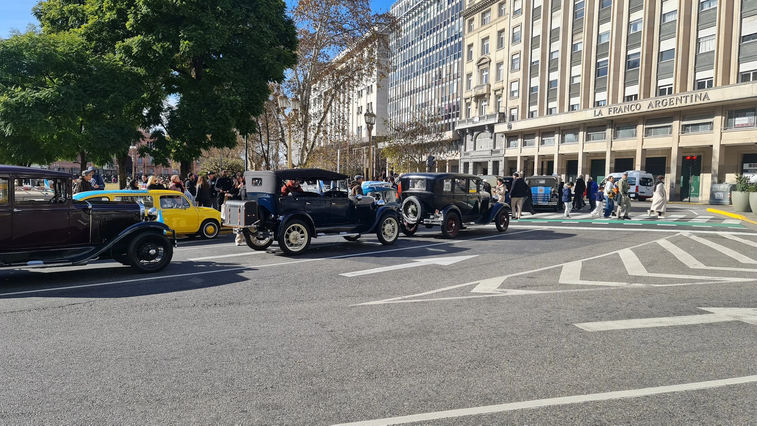 Autos antiguos en una calle de la ciudad con edificios altos y árboles, y personas caminando en la acera.