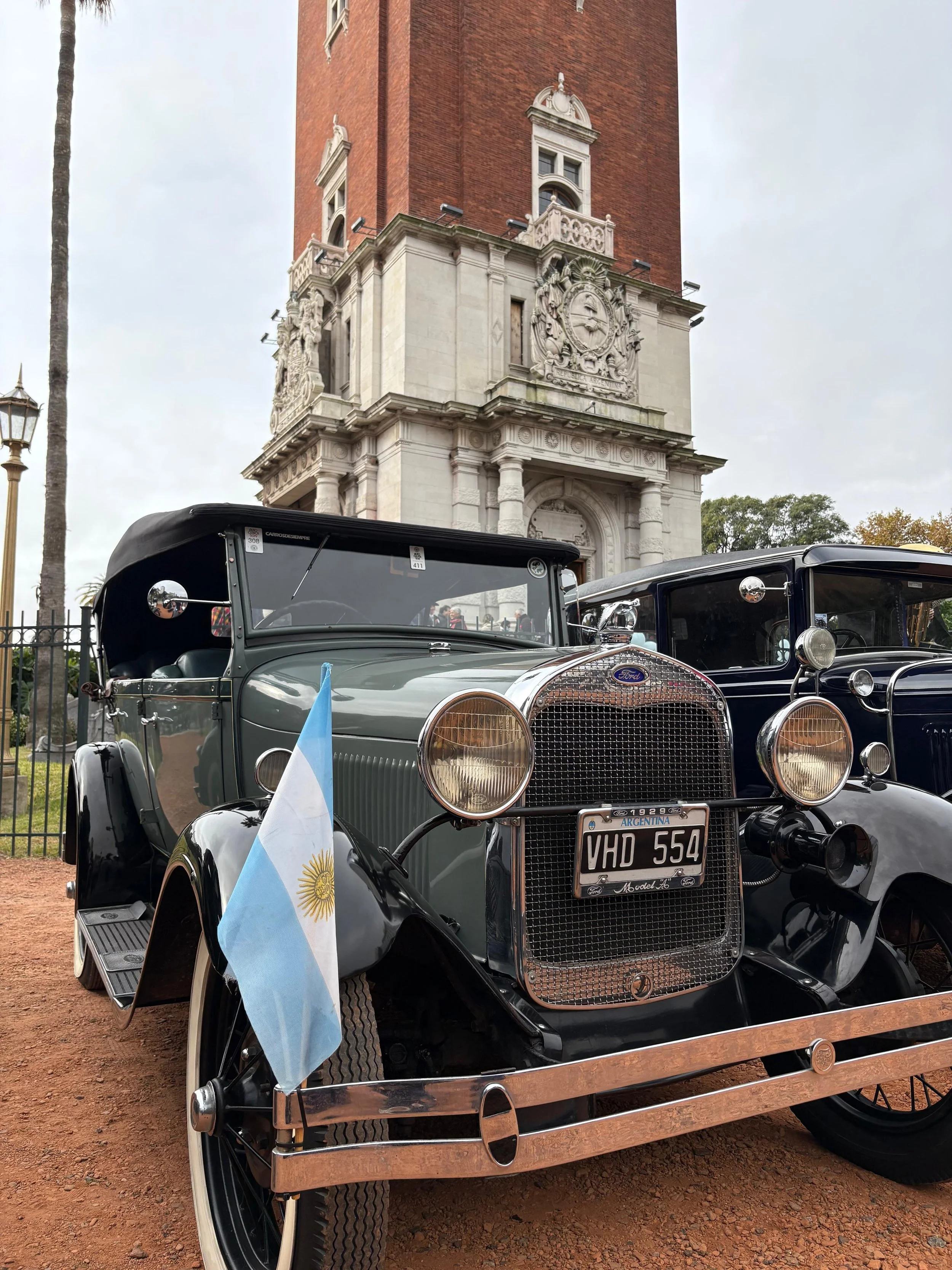 Un automóvil antiguo Ford de color gris con bandera de Argentina en el parabrisas, estacionado frente a un edificio con arquitectura clásica y un reloj en la fachada