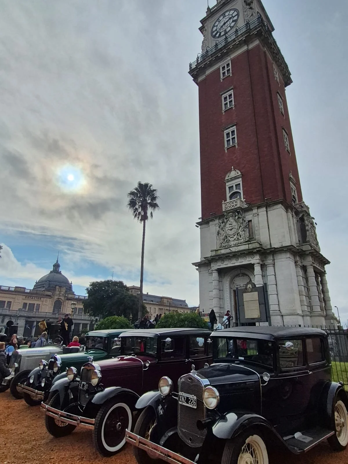 Vista de la Torre del Reloj en la ciudad con coches clásicos en primer plano, árboles y un cielo parcialmente nublado.