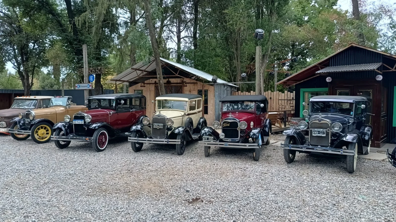 Cinco autos clásicos de diferentes colores y modelos estacionados en un área exterior con suelo de grava, rodeados de árboles y dos pequeñas estructuras de madera a los lados.